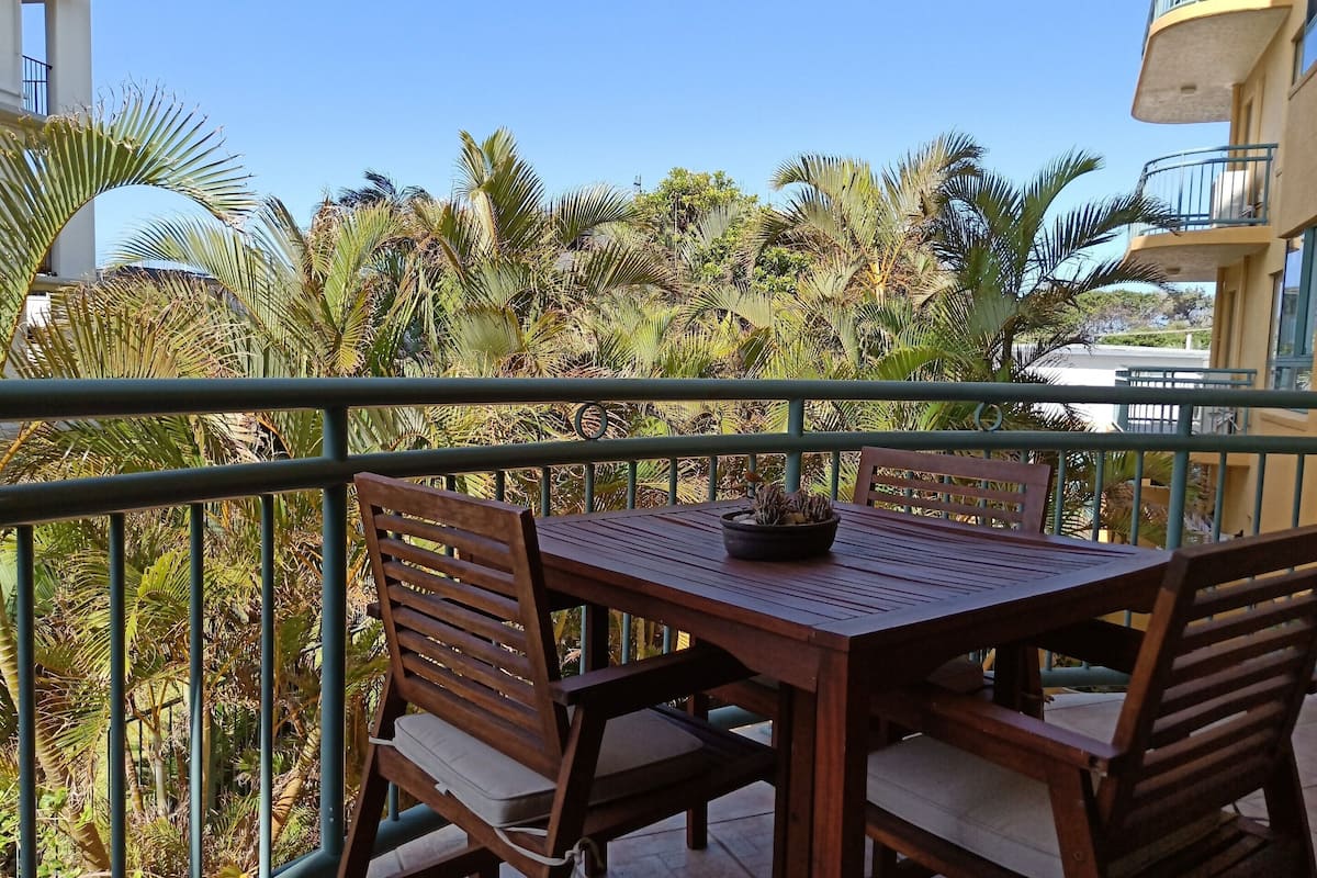 A balcony with a wooden table and chairs, surrounded by lush greenery and a clear blue sky.