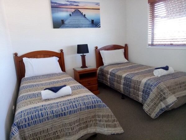 A room with two wooden beds, each with a patterned bedspread and pillows. There is a nightstand with a lamp between the beds, and a painting of a pier on the wall.