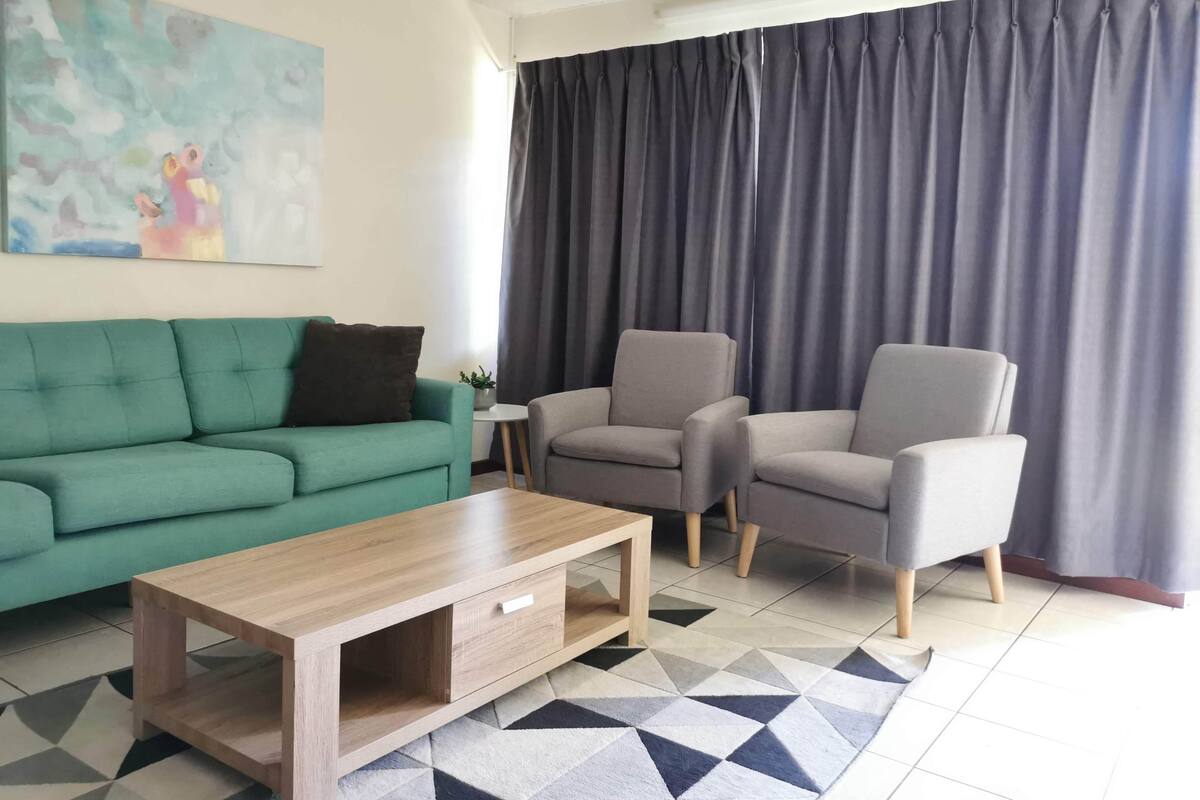 Un salon moderne avec un canapé vert, deux fauteuils gris, une table basse en bois et un tapis à motifs géométriques.