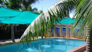 A swimming pool with a tiled edge, surrounded by palm trees and a shaded area with a green canopy. - Palm Waters Holiday Villas (North Ward)