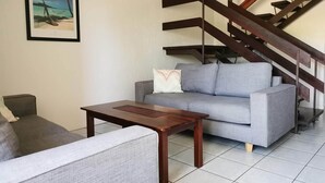 A living room with a grey sofa, a small wooden table, and a staircase with wooden railings. - Palm Waters Holiday Villas (North Ward)