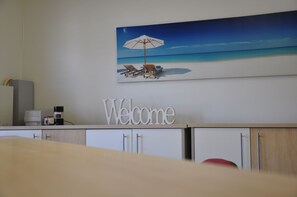 Un comptoir d’accueil avec une pancarte « Bienvenue », un tableau représentant une scène de plage et un appareil à café.