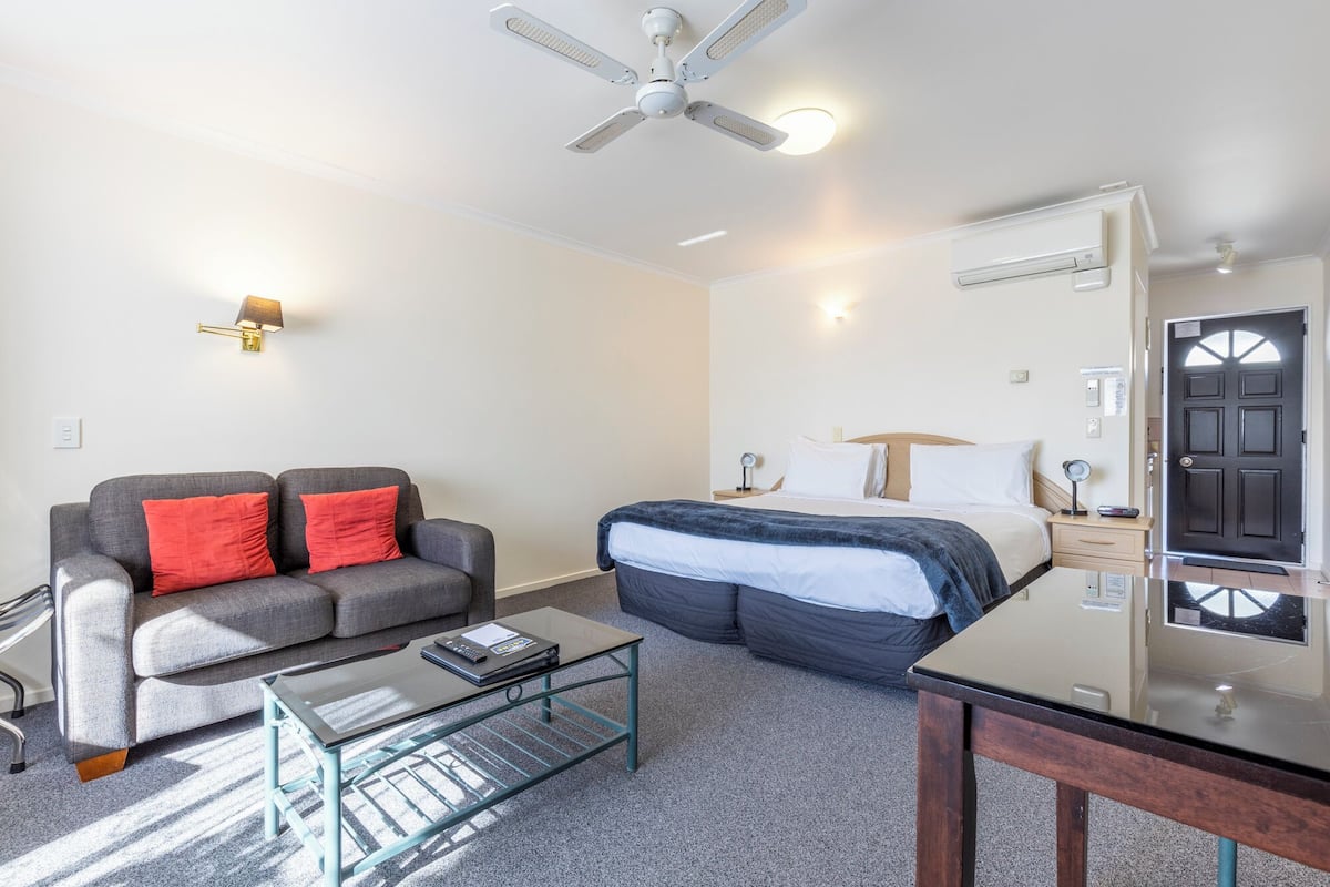 Une chambre d’hôtel moderne avec un canapé gris, un lit recouvert d’une couverture bleue, une table basse en verre et un ventilateur de plafond.
