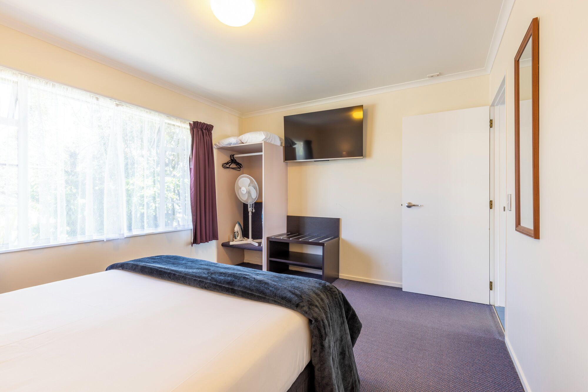 A hotel room with a bed, a desk with a chair, a wardrobe, and a TV mounted on the wall.