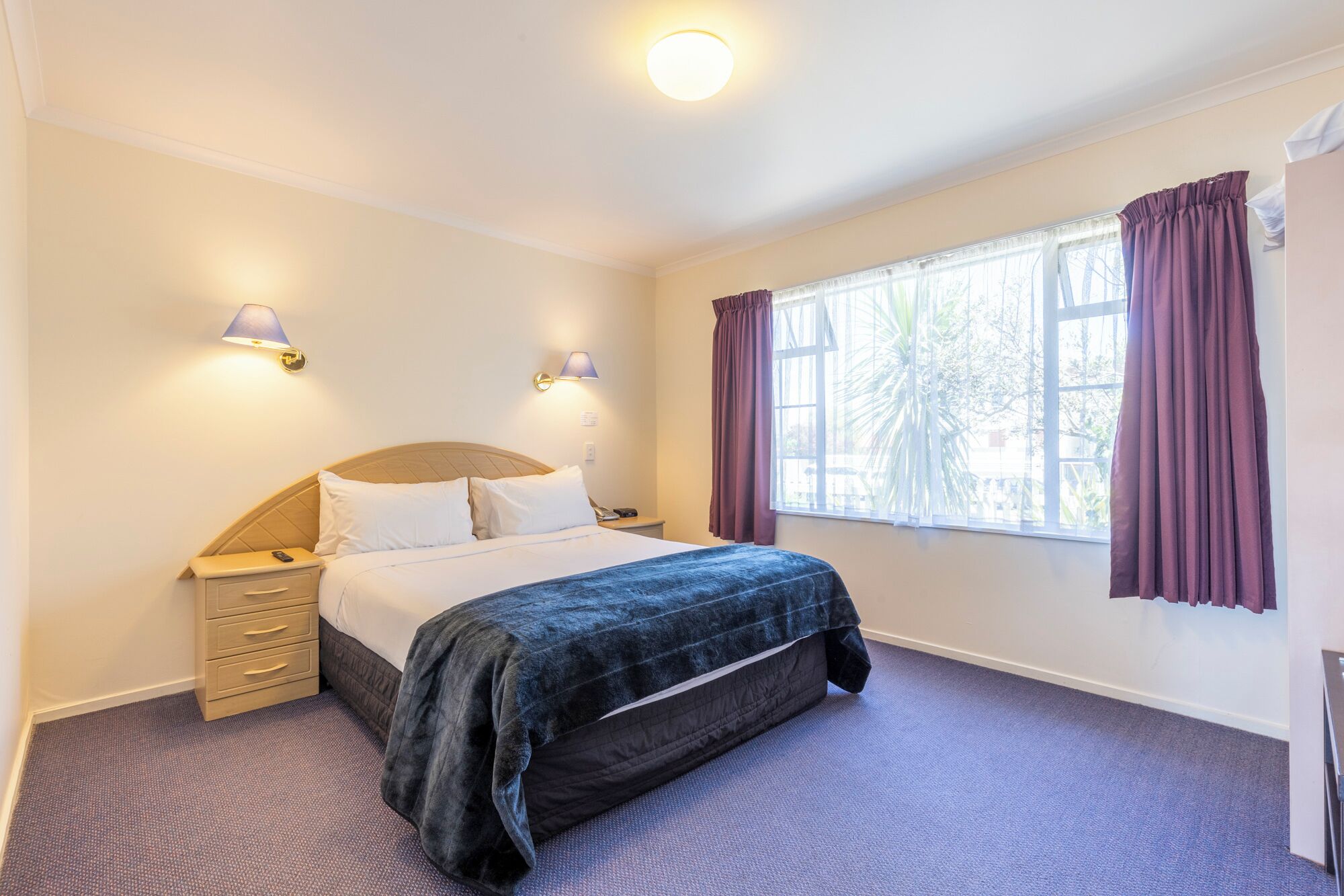 A bedroom with a bed, bedside tables, two wall-mounted lamps, a window with curtains, and a carpeted floor.
