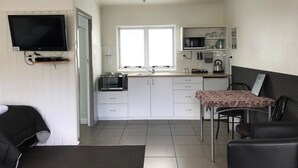 Une cuisine compacte avec des meubles blancs, un four à micro-ondes et une bouilloire. On y trouve une table à manger avec des chaises et un téléviseur fixé au mur.