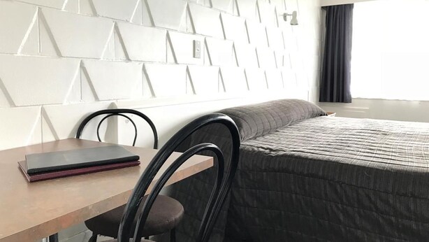 Une chambre d’hôtel moderne avec une tête de lit à motifs géométriques, un lit recouvert d’une courtepointe, un bureau en bois avec un carnet et une chaise.