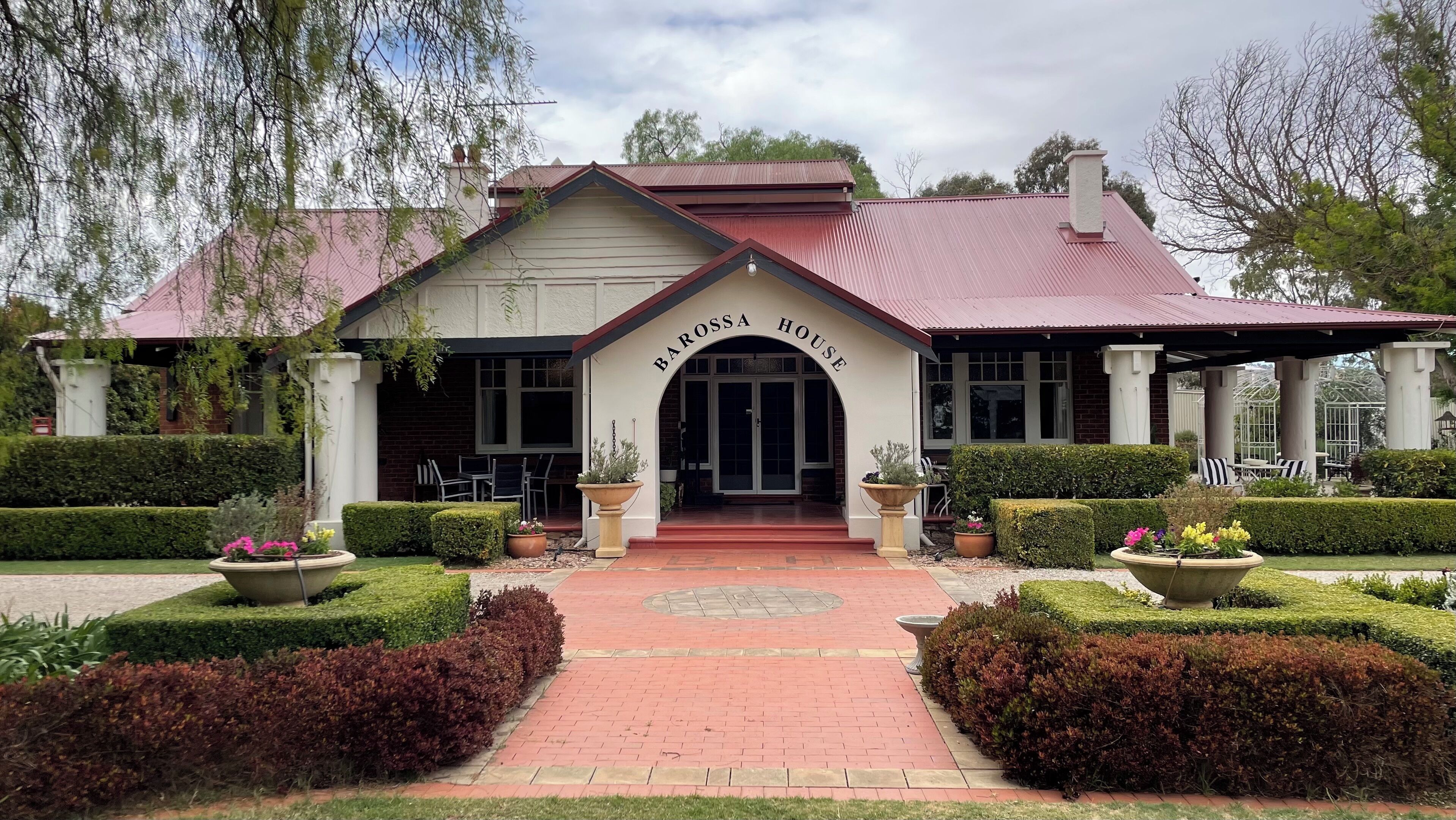 Una casa en Barossa con techo rojo, una entrada central y un jardín bien cuidado con setos y plantas en macetas.