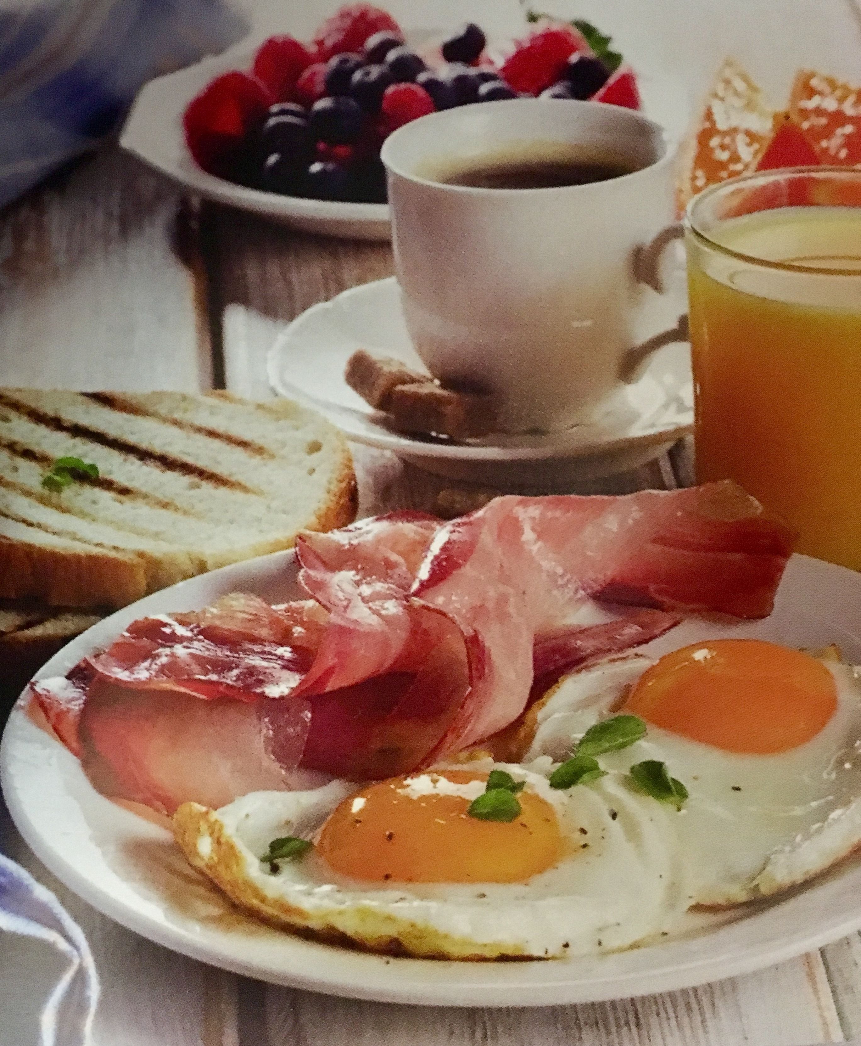 Un plato de desayuno con huevos, prosciutto, tostadas, una taza de café y un tazón de frutos rojos.
