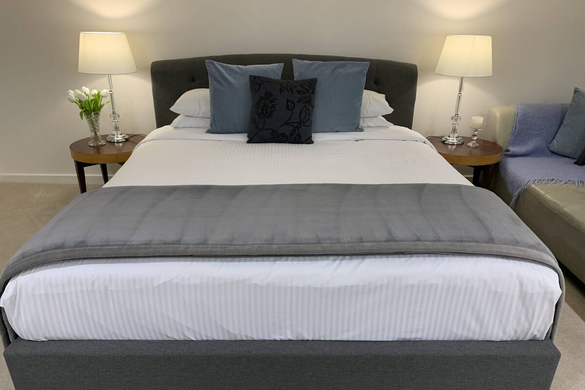 A bedroom with a large bed, two bedside tables with lamps, a nightstand, a sofa, and a vase with flowers.