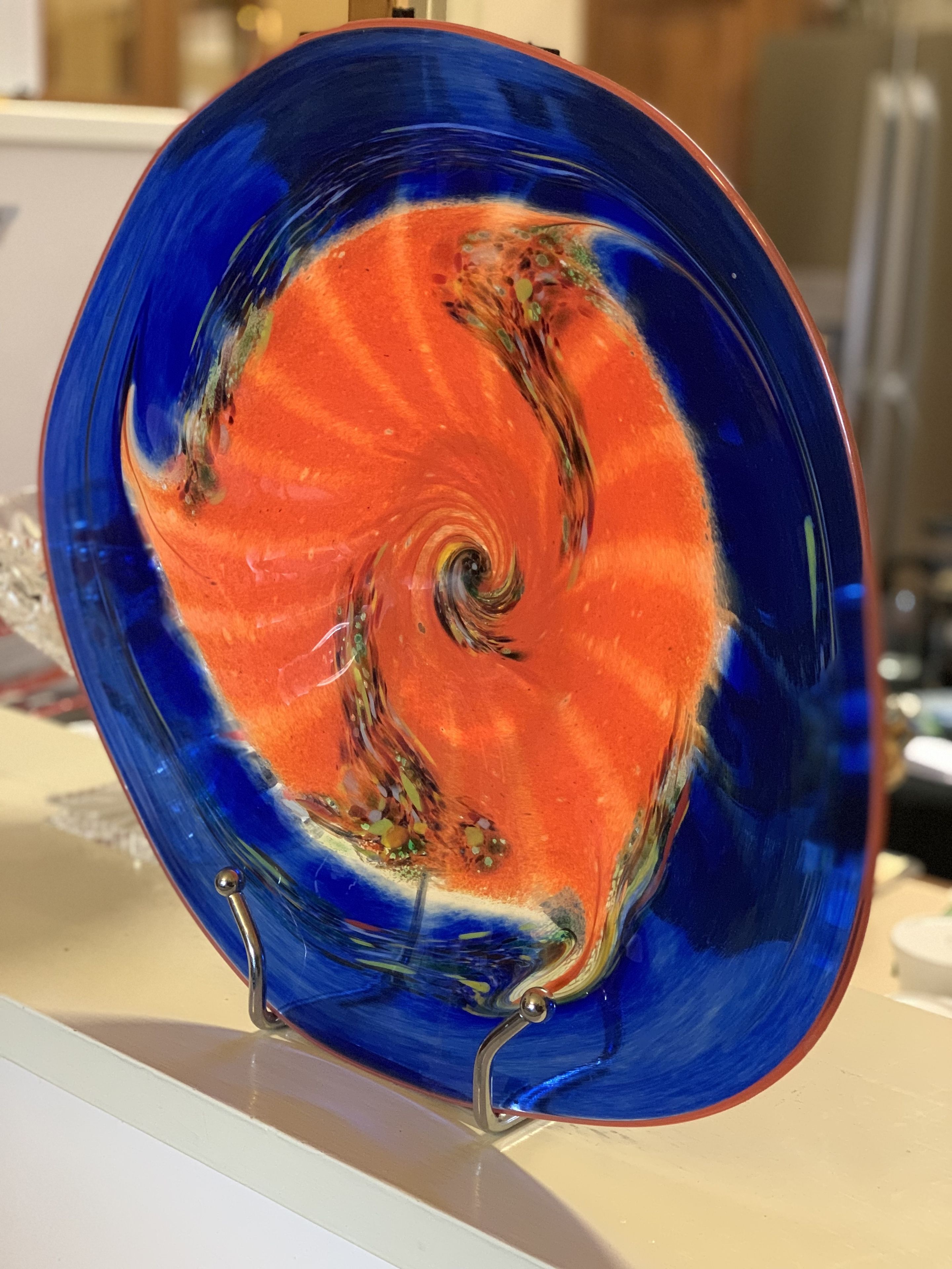 Un plato de vidrio azul y naranja con un diseño en espiral, exhibido sobre un pedestal.