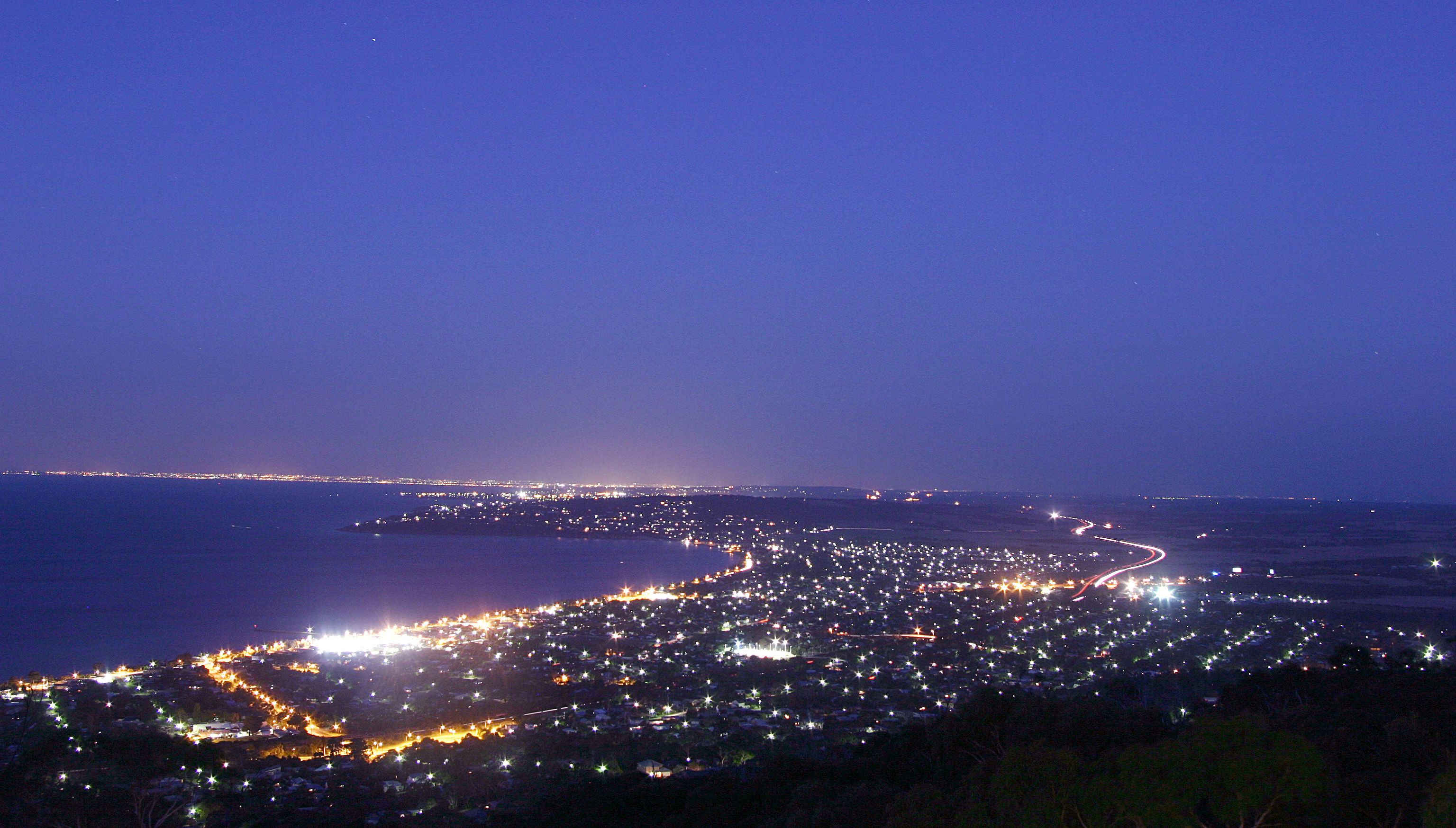 A coastal cityscape at night with a winding road along the coastline and a view of the sea.
