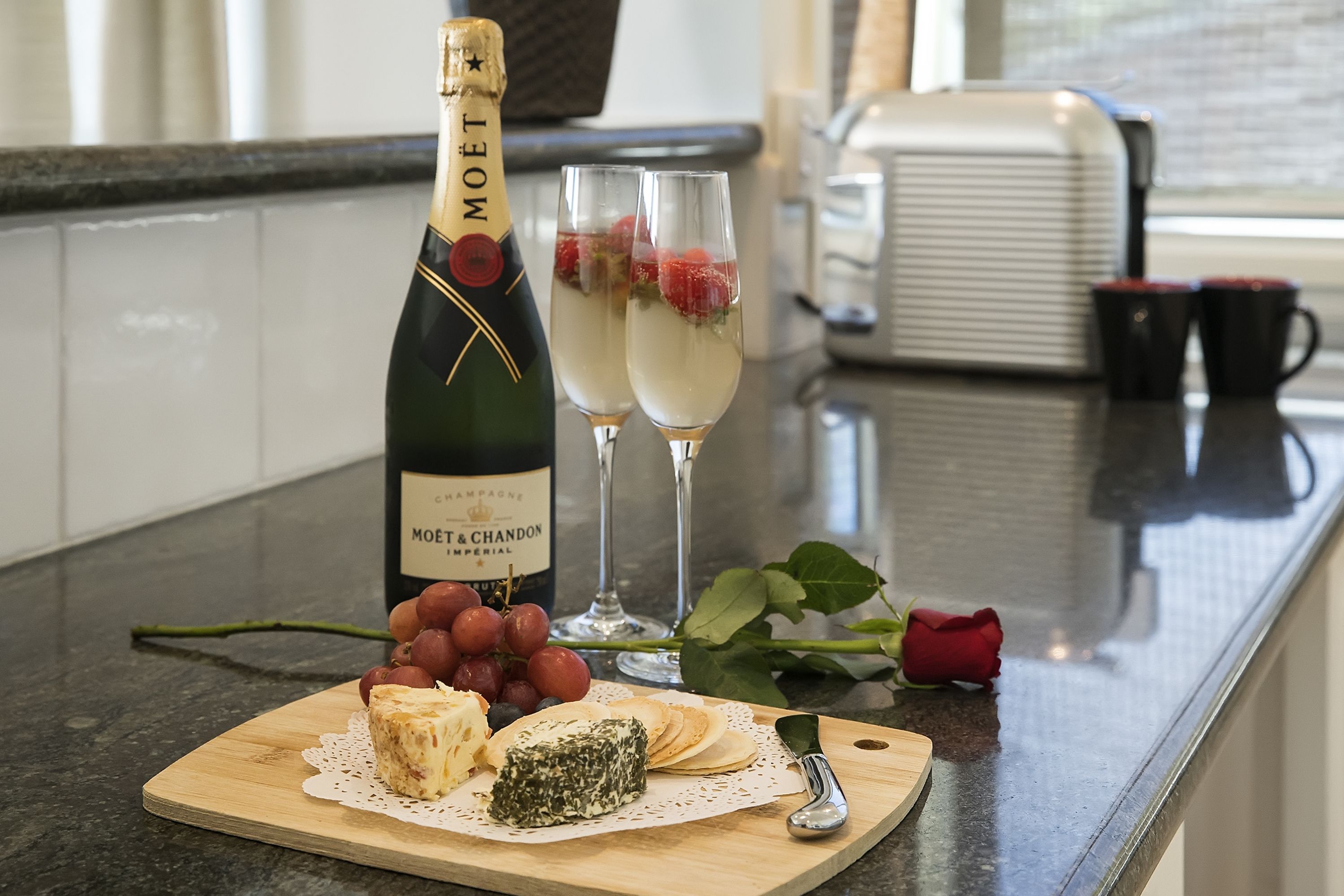Una encimera de cocina con una botella de Moët & Chandon, dos copas de champán, una variedad de quesos, uvas y una rosa roja.