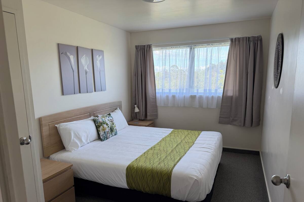 A hotel room with a bed, a nightstand, a lamp, a window with curtains, and wall art.