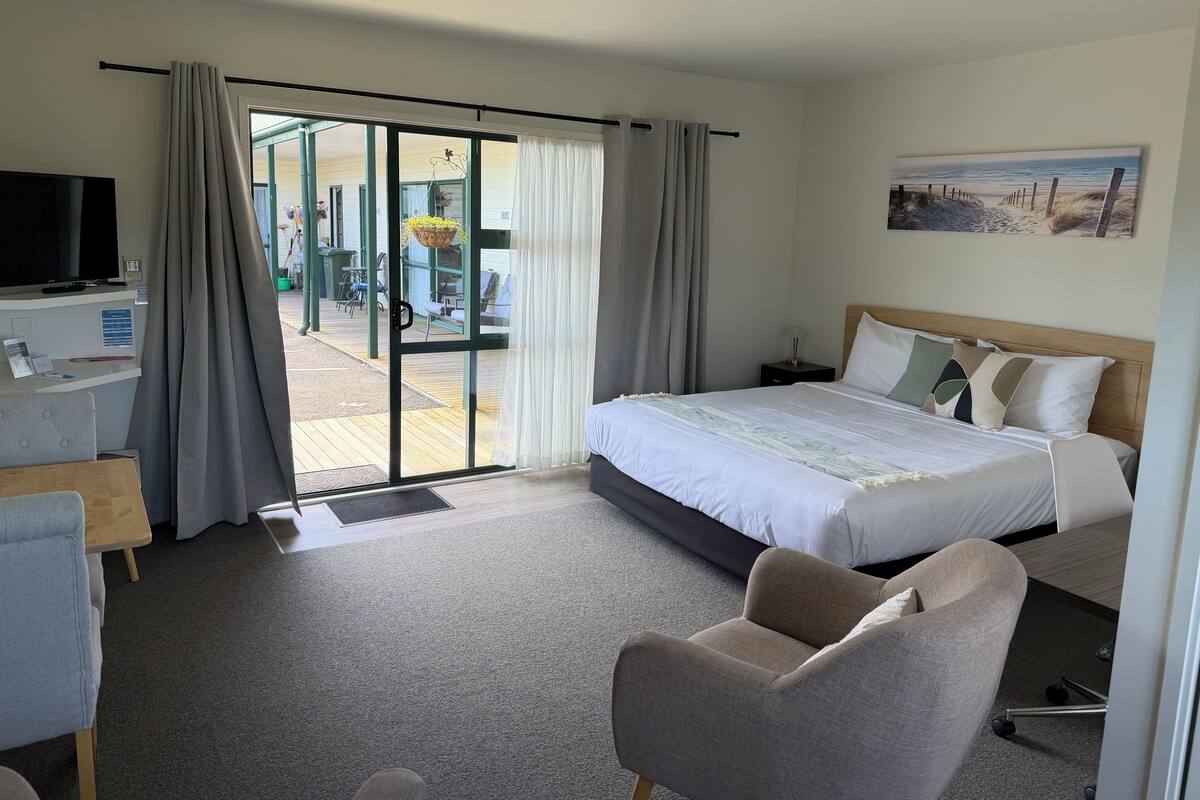 A hotel room with a bed, a desk with a TV, a chair, and a view of the outside.