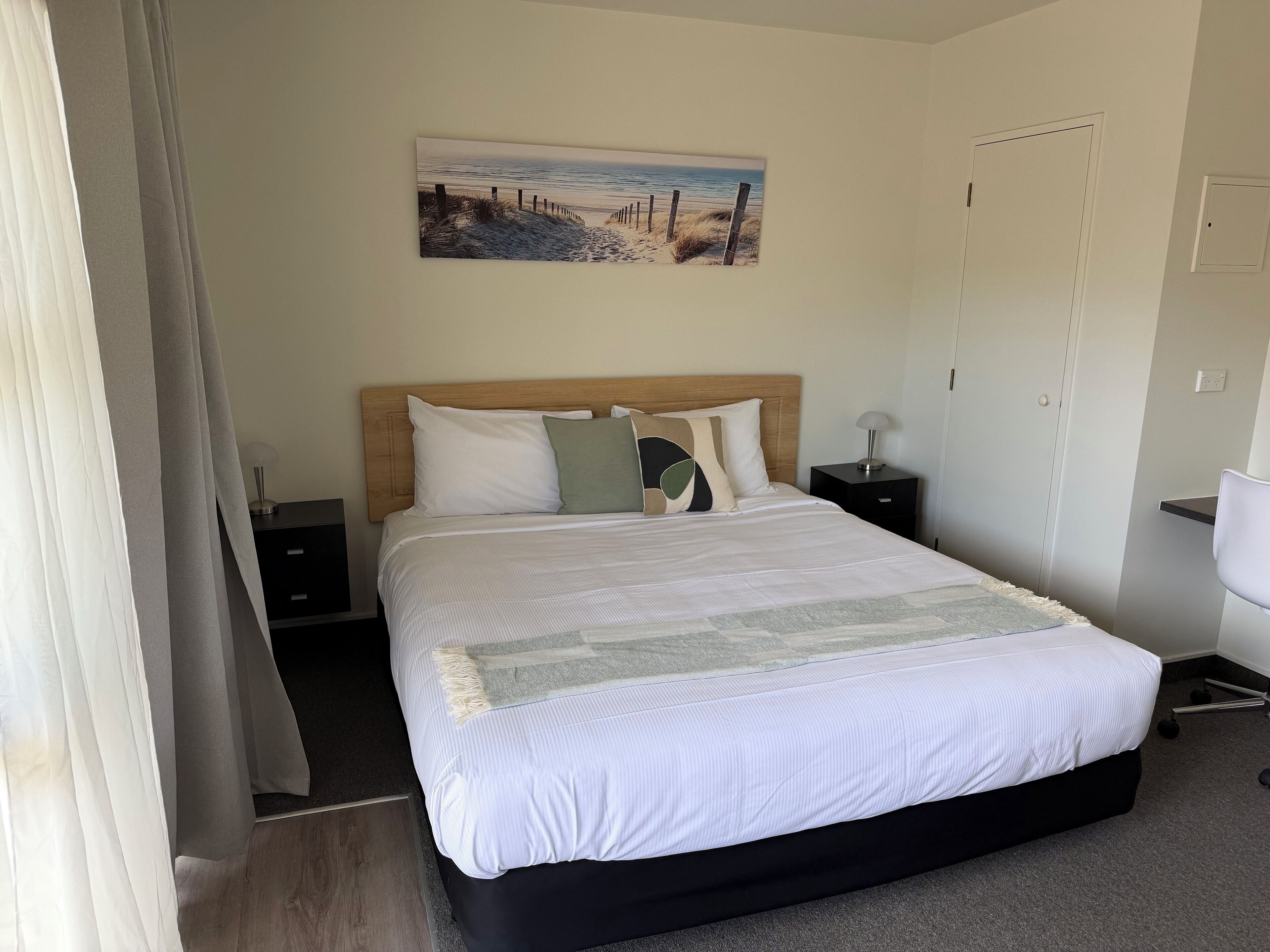 A neatly made bed with a headboard, a bedside table with a lamp, and a painting of a beach scene on the wall.
