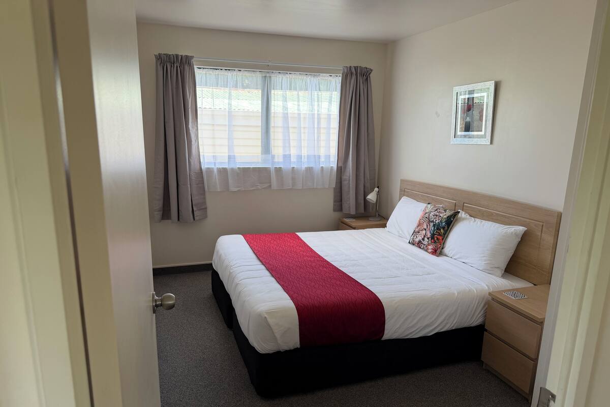 A neatly arranged hotel room with a large bed, a bedside table, a nightstand, a window with curtains, and a framed picture on the wall.
