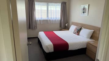 Une chambre d’hôtel bien rangée, avec un grand lit, une table de chevet, une commode, une fenêtre avec des rideaux et un tableau encadré au mur.