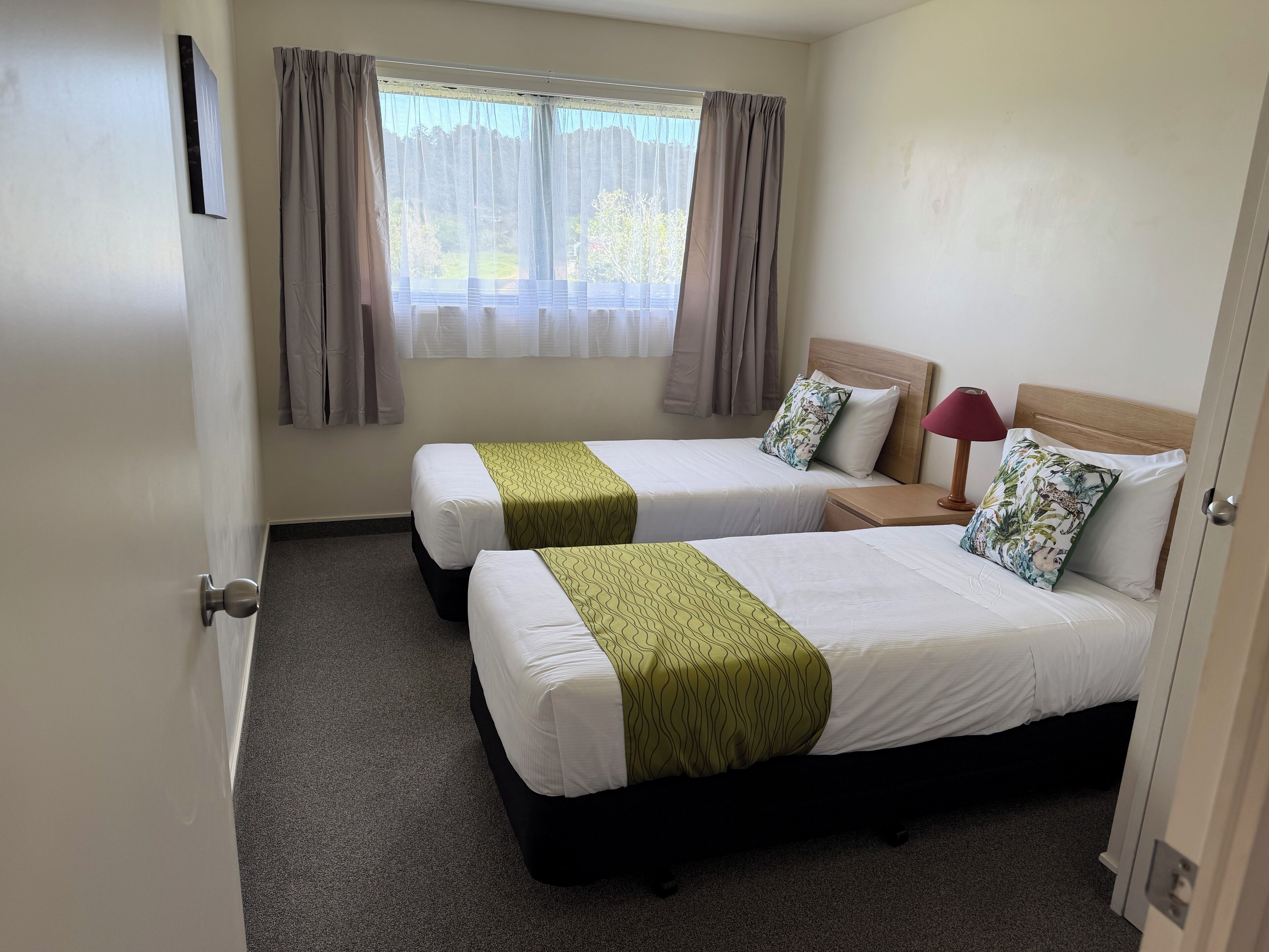 A hotel room with two beds, a window with curtains, a bedside lamp, and a door.
