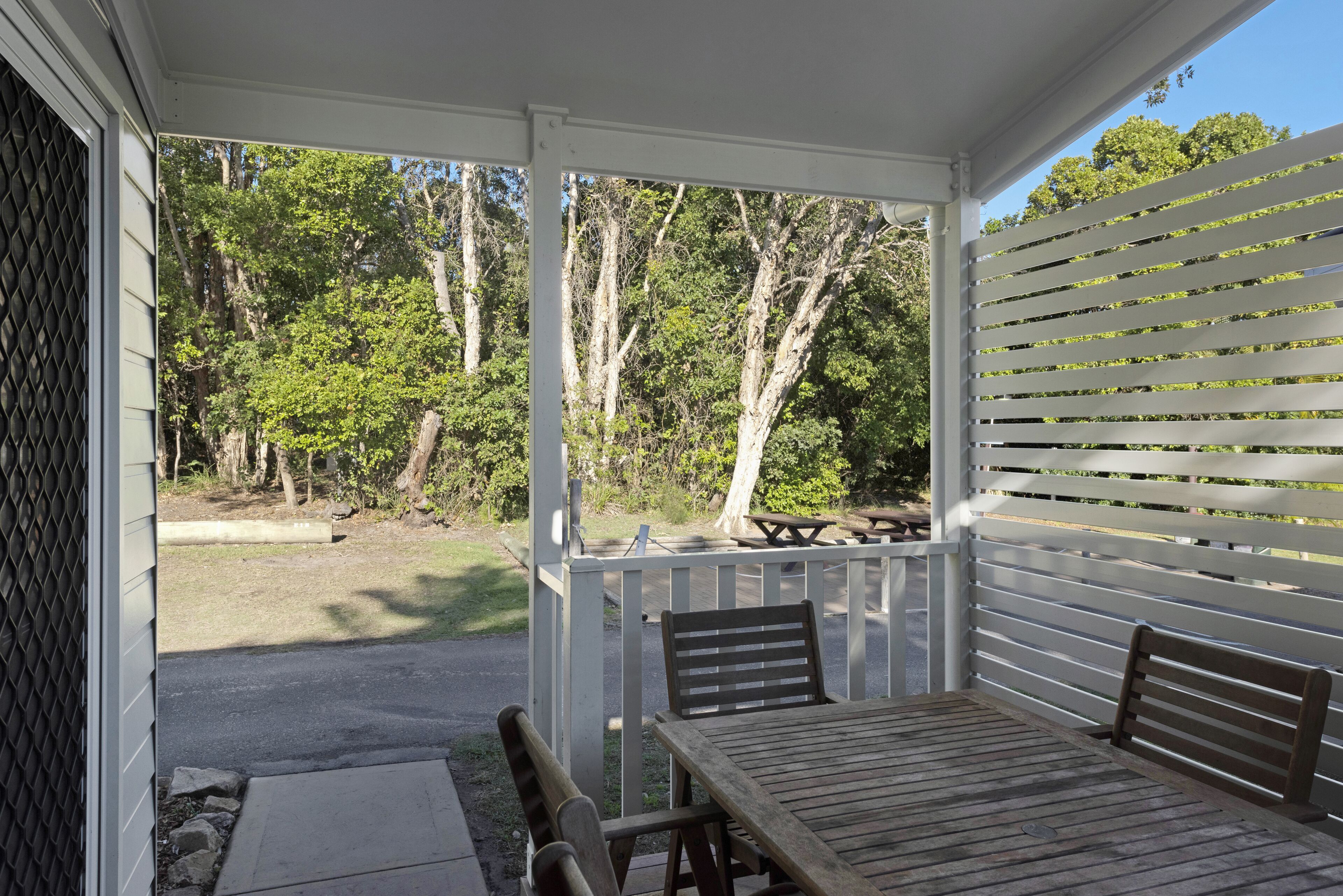 Une terrasse couverte avec une table et des chaises en bois, une table de pique-nique et une vue sur une zone boisée.