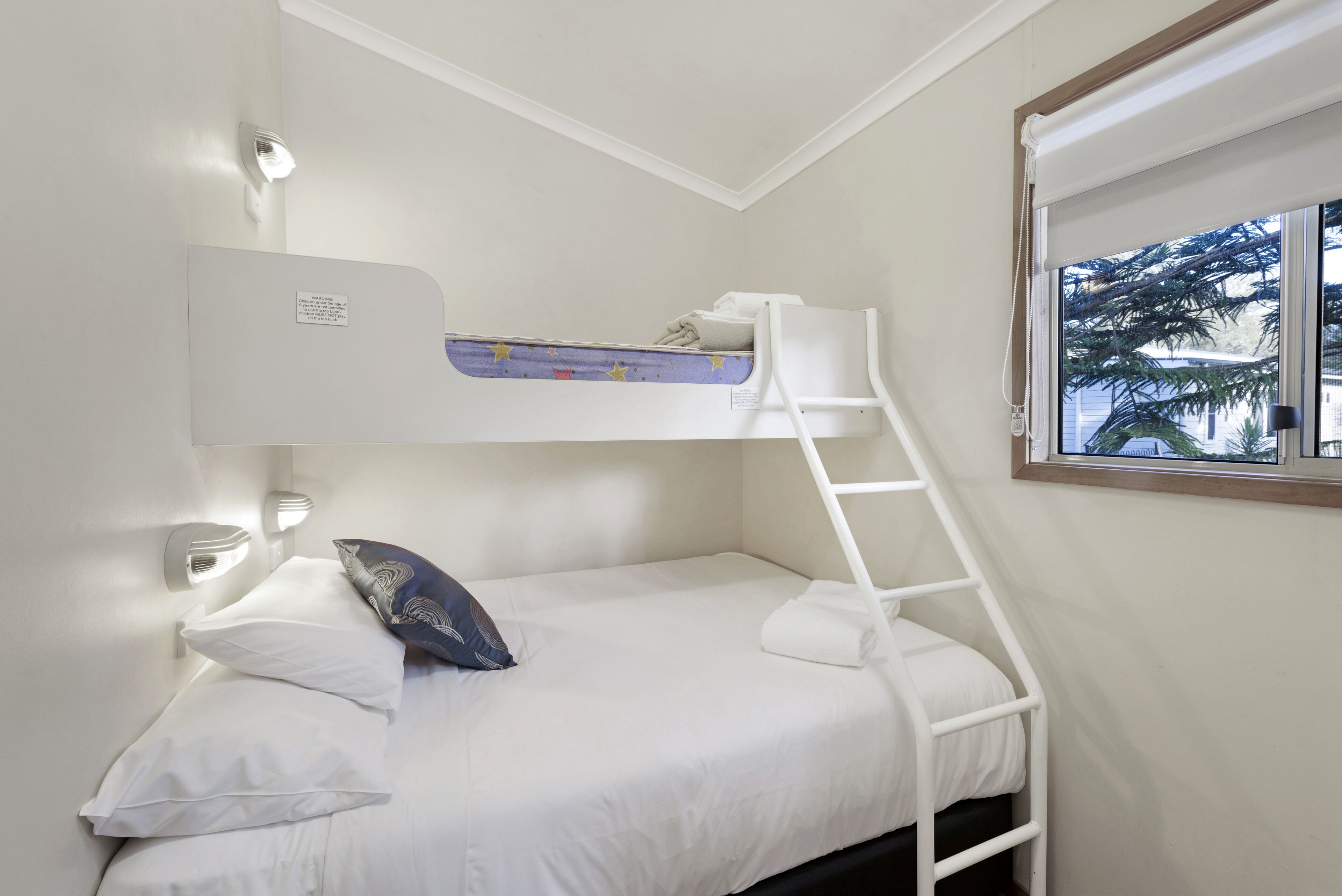 A bunk bed with a ladder, white bedding, and a window with a view of palm trees.