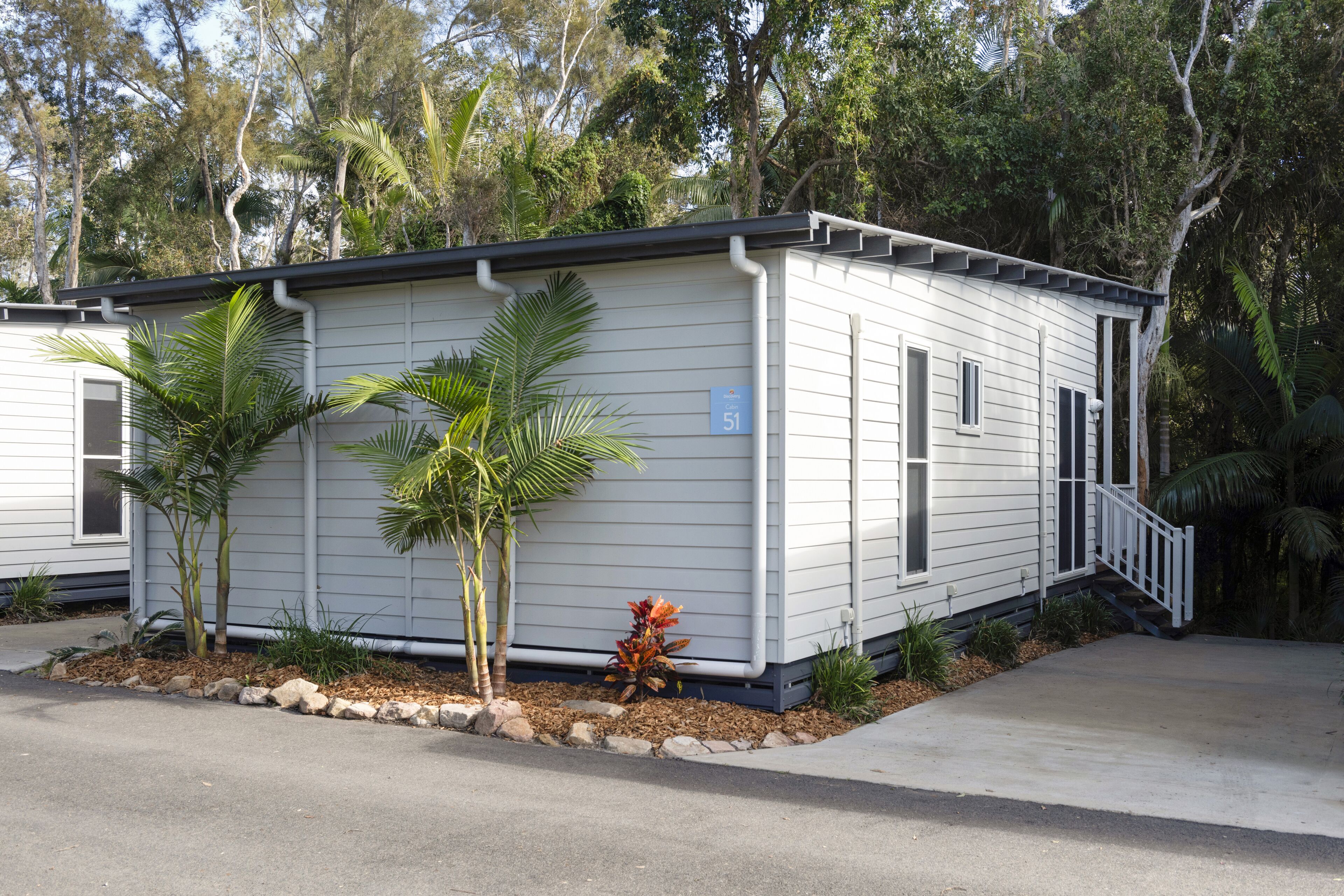 A single-story, white mobile home with a number 51 sign, surrounded by a small garden with palm trees and shrubs.