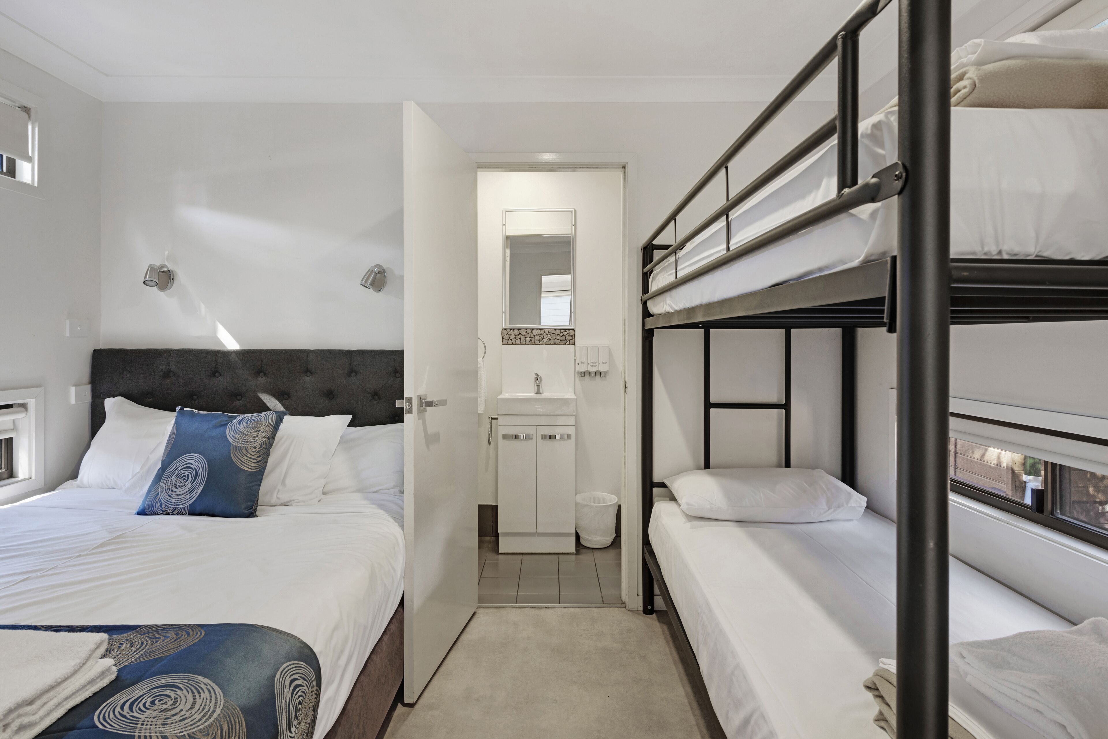 A hotel room with a bunk bed, a single bed, a bathroom with a mirror, and a small window.