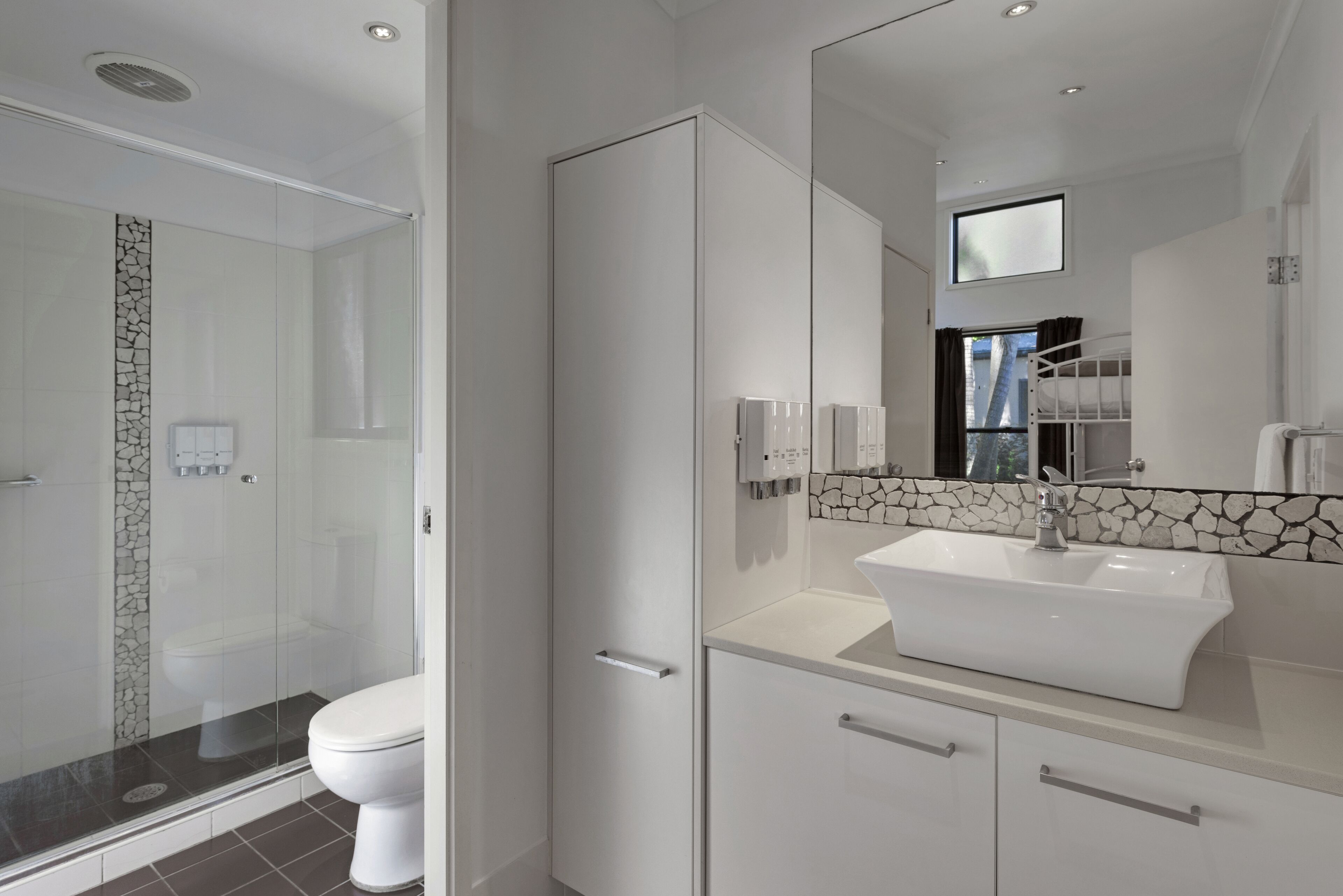 Ein modernes Badezimmer mit einer gläsernen Duschkabine, einer weißen Toilette, einem weißen Waschbecken mit Steinrückwand und einem großen Spiegel.