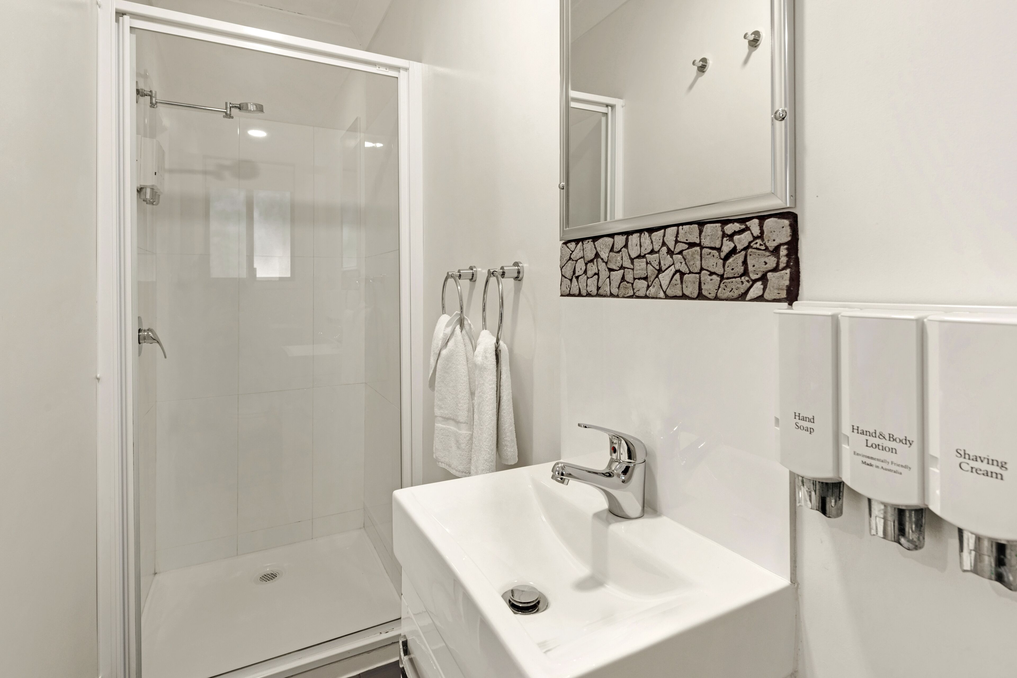 A modern bathroom with a glass shower enclosure, a white sink, and a mirror above it. There are towels hanging on a rack and a soap dispenser on the wall.