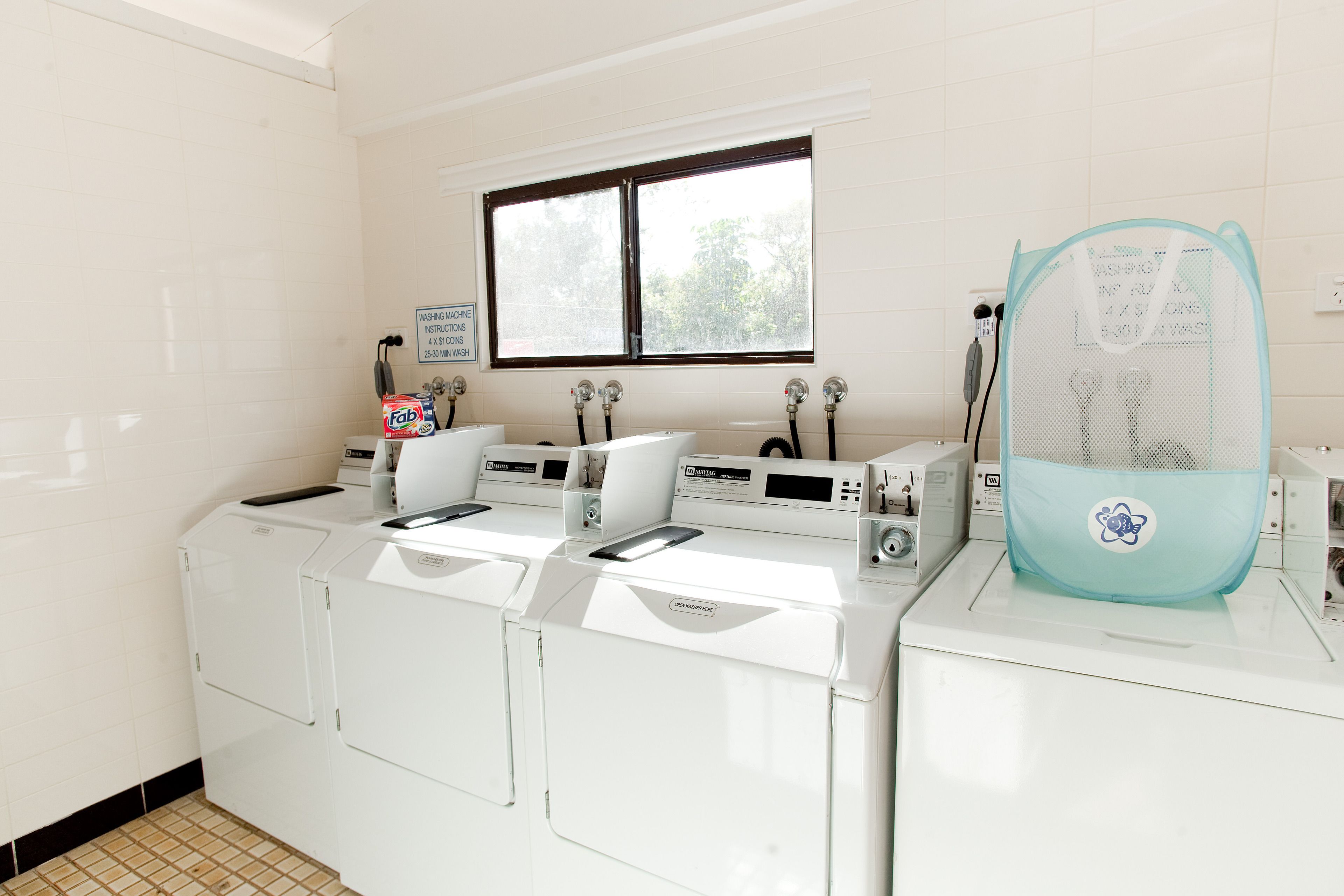 A laundry area with four washing machines, a window, and a blue laundry basket.