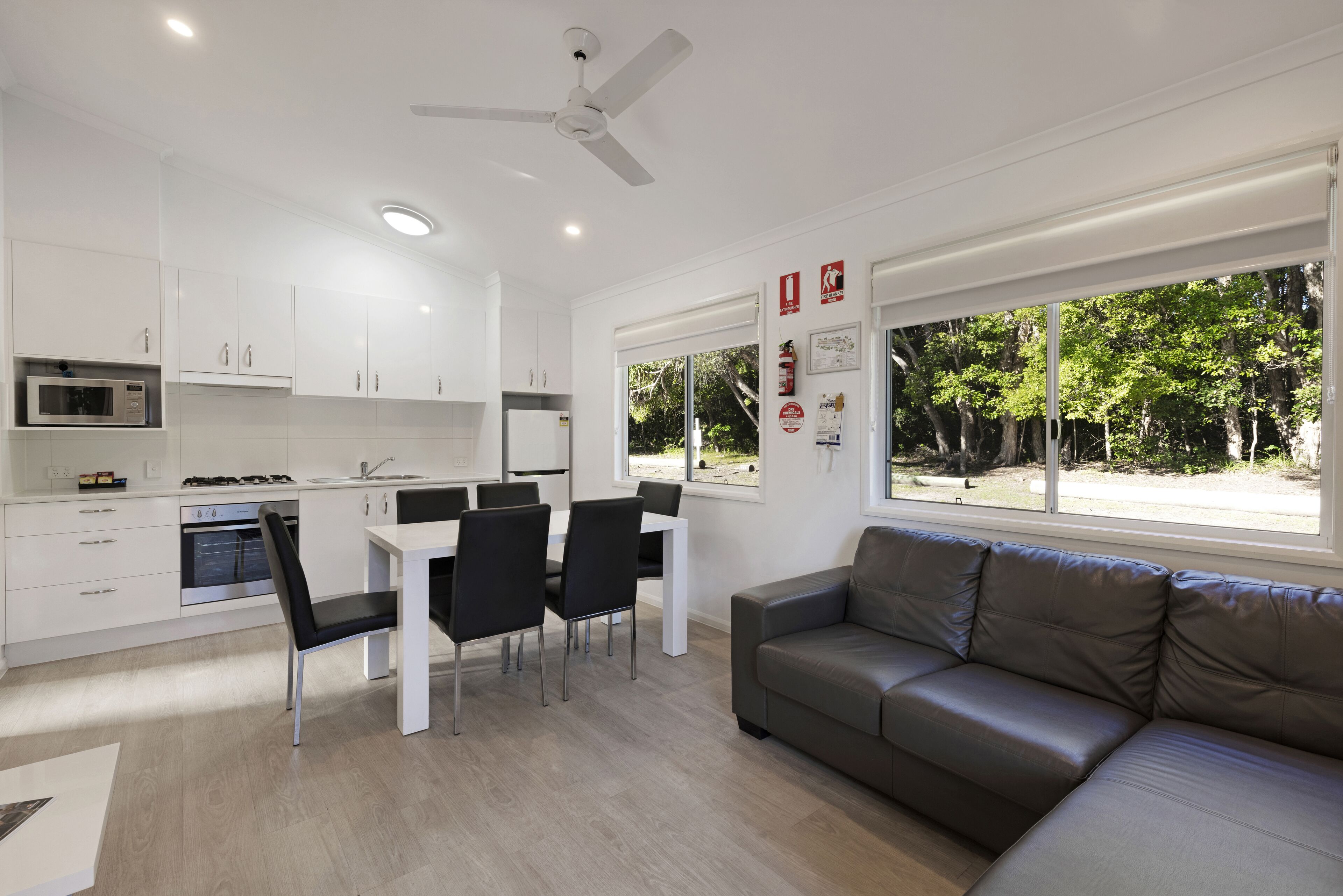 A modern kitchen with white cabinets, a dining area with a white table and black chairs, and a grey sofa near large windows with a view of trees.