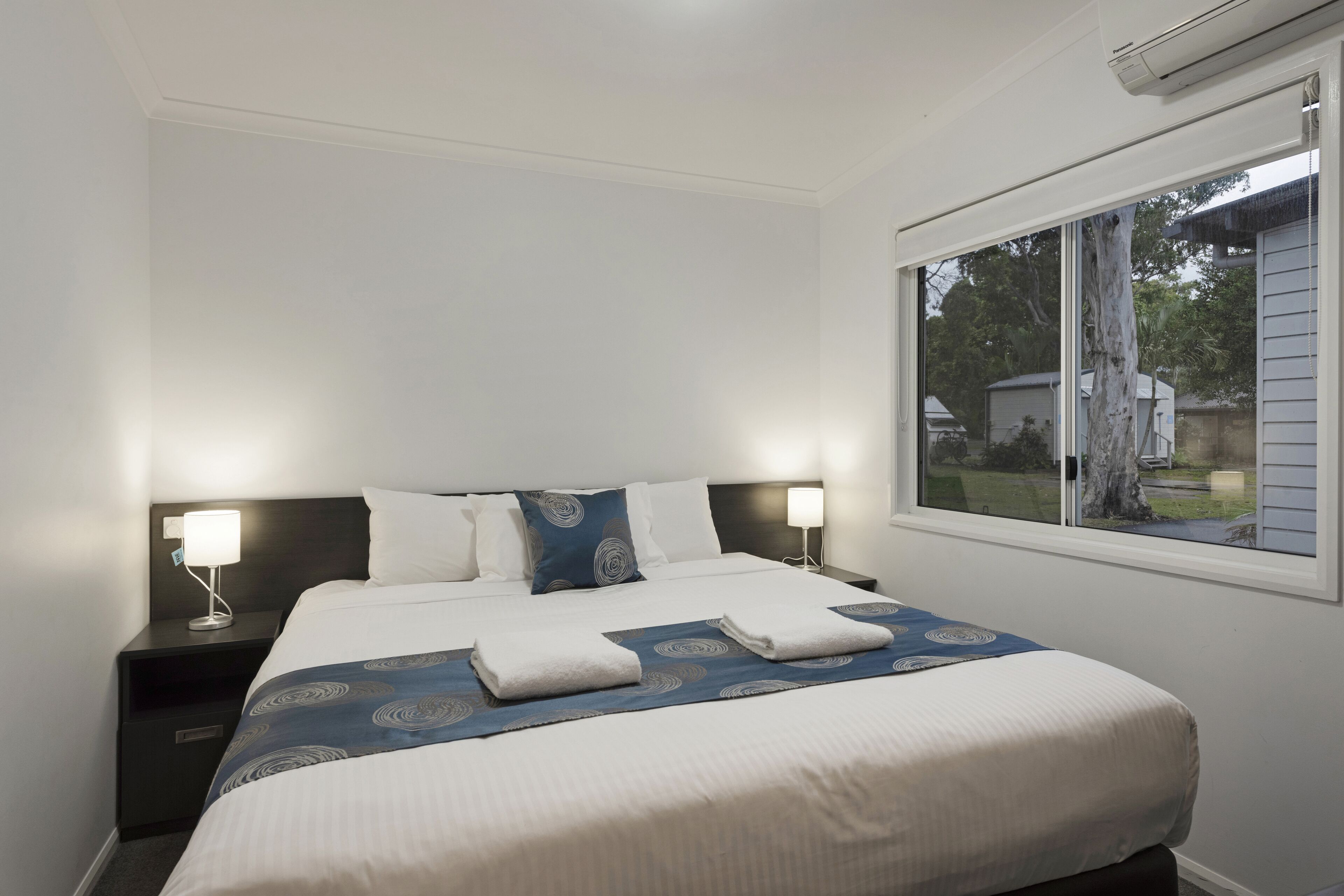 Ein ordentlich bezogenes Bett mit einer blauen und weißen Bettdecke, zwei Kissen und zwei gefaltete Handtücher. Ein Nachttisch mit Lampe und ein Fenster mit Blick ins Freie sind ebenfalls zu sehen.