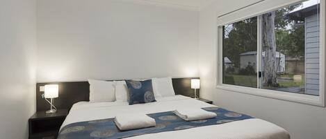 Ein ordentlich bezogenes Bett mit einer blauen und weißen Bettdecke, zwei Kissen und zwei gefaltete Handtücher. Ein Nachttisch mit Lampe und ein Fenster mit Blick ins Freie sind ebenfalls zu sehen.