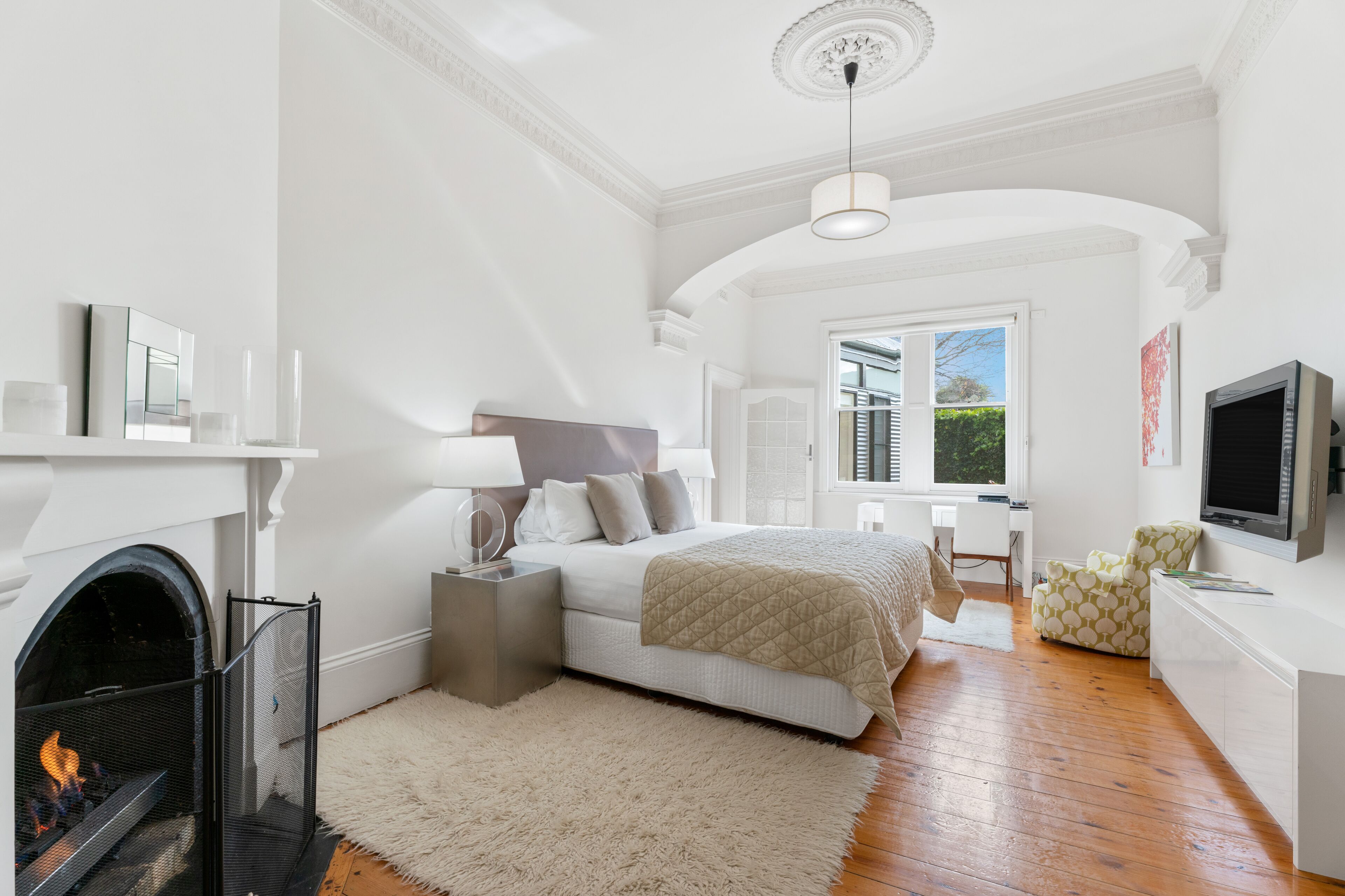 A bedroom with a fireplace, a bed with a quilted blanket, a desk, a chair, and a television.