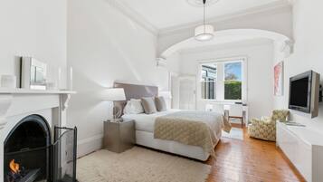 A bedroom with a fireplace, a bed with a quilted blanket, a desk, a chair, and a television.