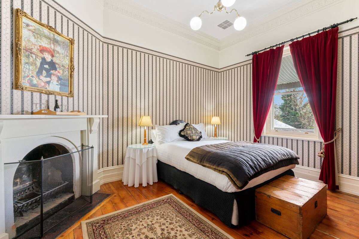 Un dormitorio con chimenea, una cama con cabecera oscura, dos mesitas de noche con lámparas y una ventana con cortinas rojas.