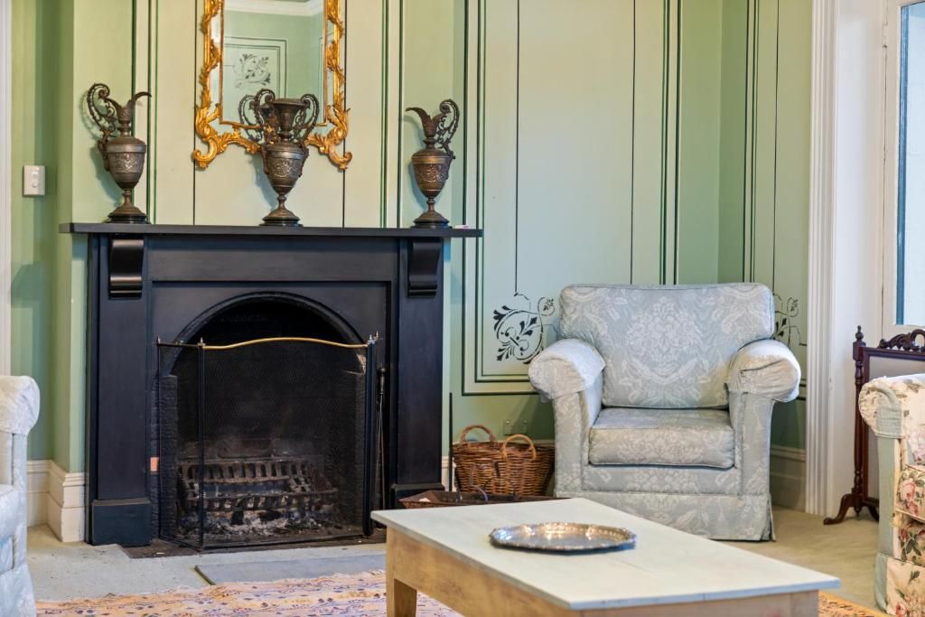 A room with a fireplace, a patterned armchair, a small table, and a mirror with decorative vases.