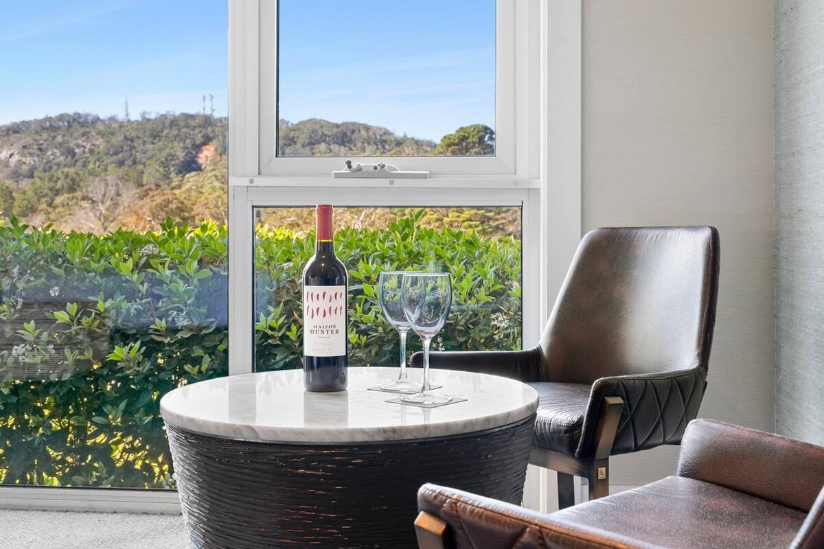 A round table with a bottle of wine and a glass, a leather chair, and a view of greenery through a window.