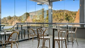 A modern outdoor seating area with wicker chairs and tables, large glass windows offering a view of a lake and forested hills. - The Gibraltar  (Bowral)