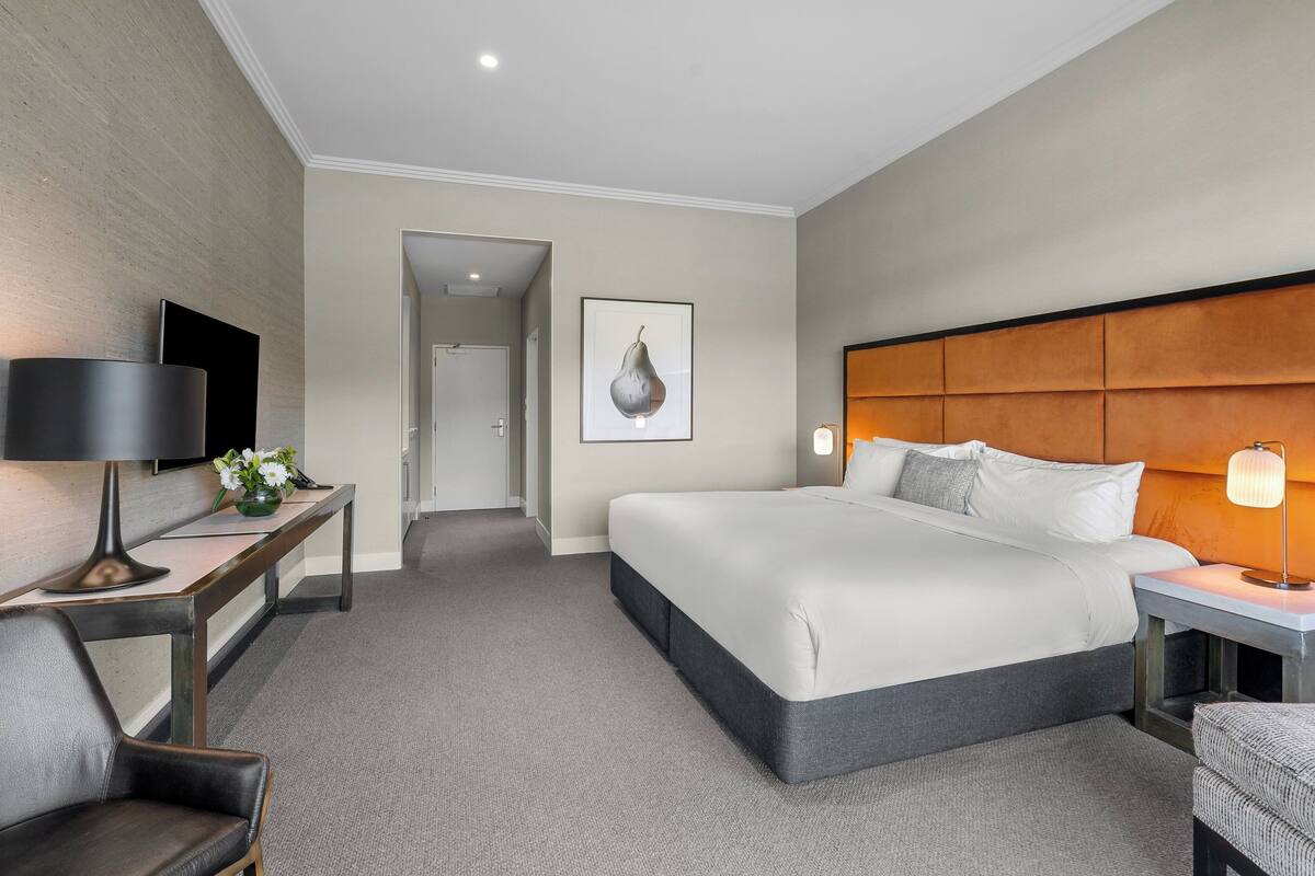 A modern hotel room with a large bed, a desk with a lamp, a TV, and a framed artwork on the wall.