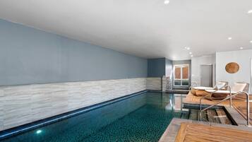 A modern indoor swimming pool with clear water, a wooden deck, and lounge chairs.