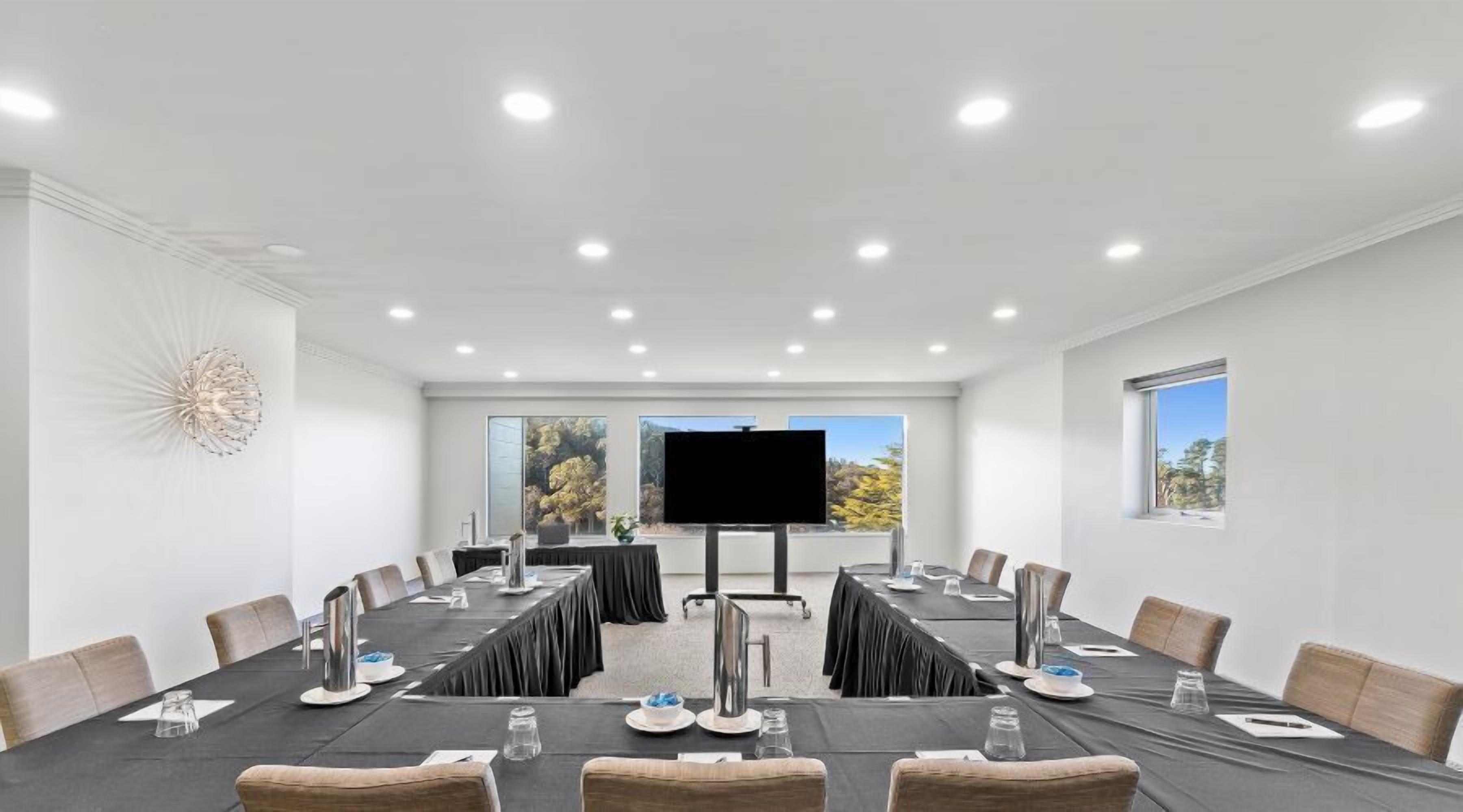 A conference room with a long table, chairs, a large screen, and windows with a view of trees.