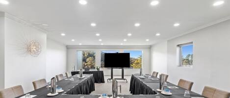 A conference room with a long table, chairs, a large screen, and windows with a view of trees.