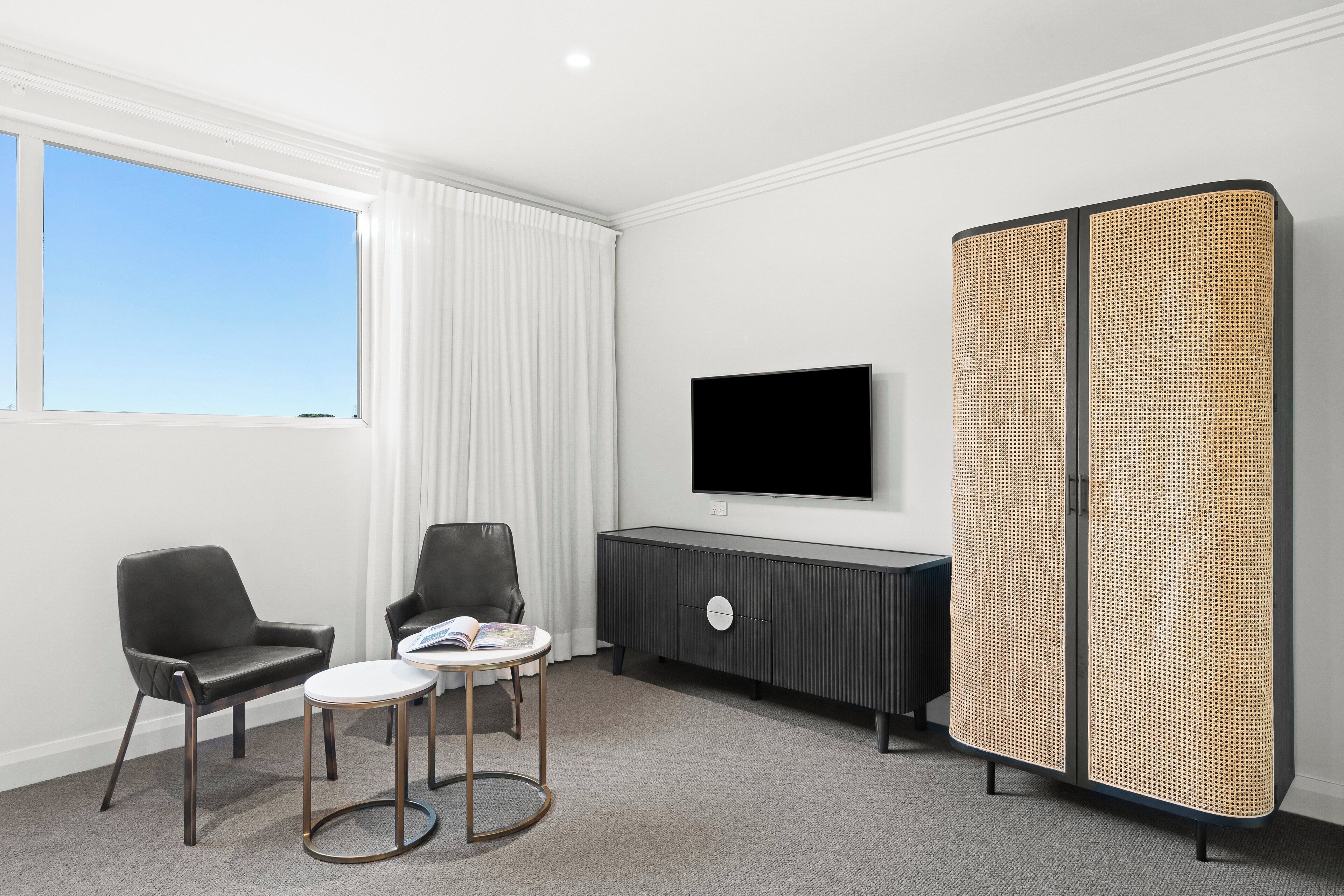 Una habitación de hotel moderna con un ventanal grande, un televisor de pantalla plana, un sofá de cuero negro, una mesita redonda y un separador tejido.