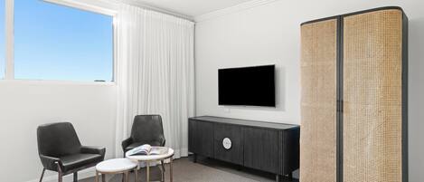 A modern hotel room with a large window, a flat-screen TV, a black leather sofa, a small round table, and a woven partition.