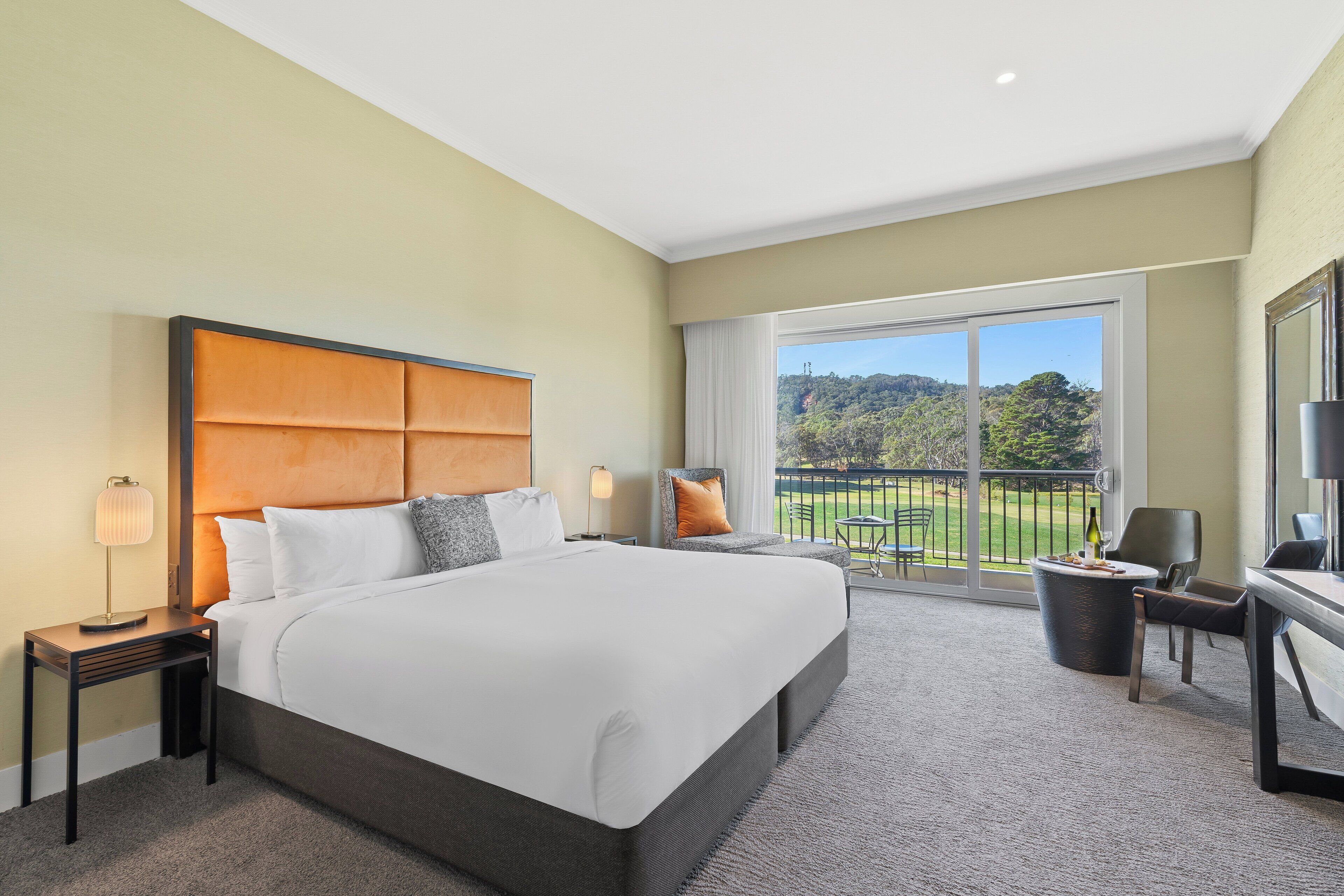 A hotel room with a large bed, a balcony with a view of the greenery, and a seating area with a table and chairs.