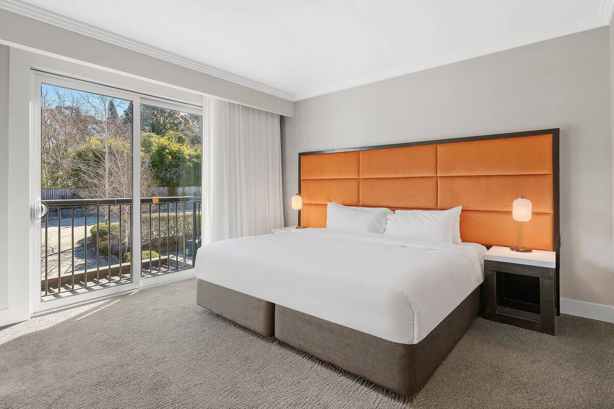 A hotel room with a large bed, a nightstand, a lamp, and a view of the outdoors through a sliding door.
