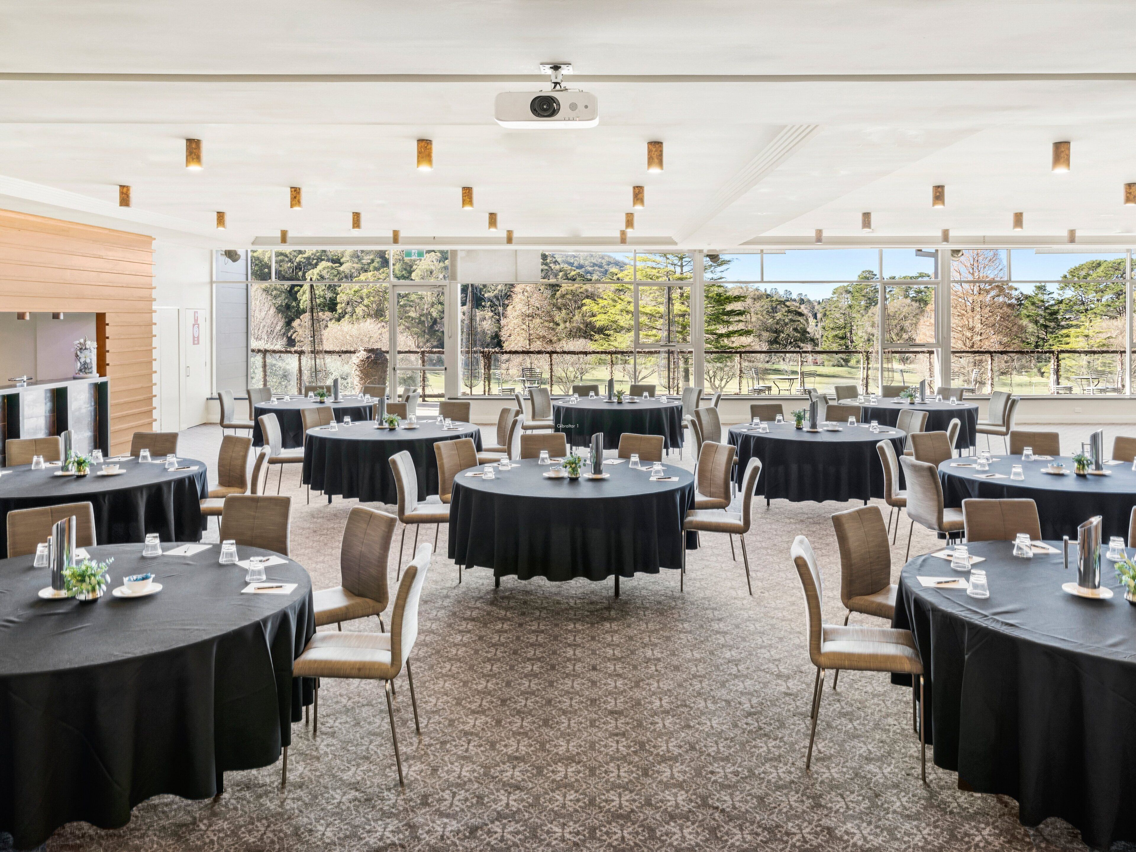 A conference room with multiple round tables, chairs, and a projector on the ceiling.