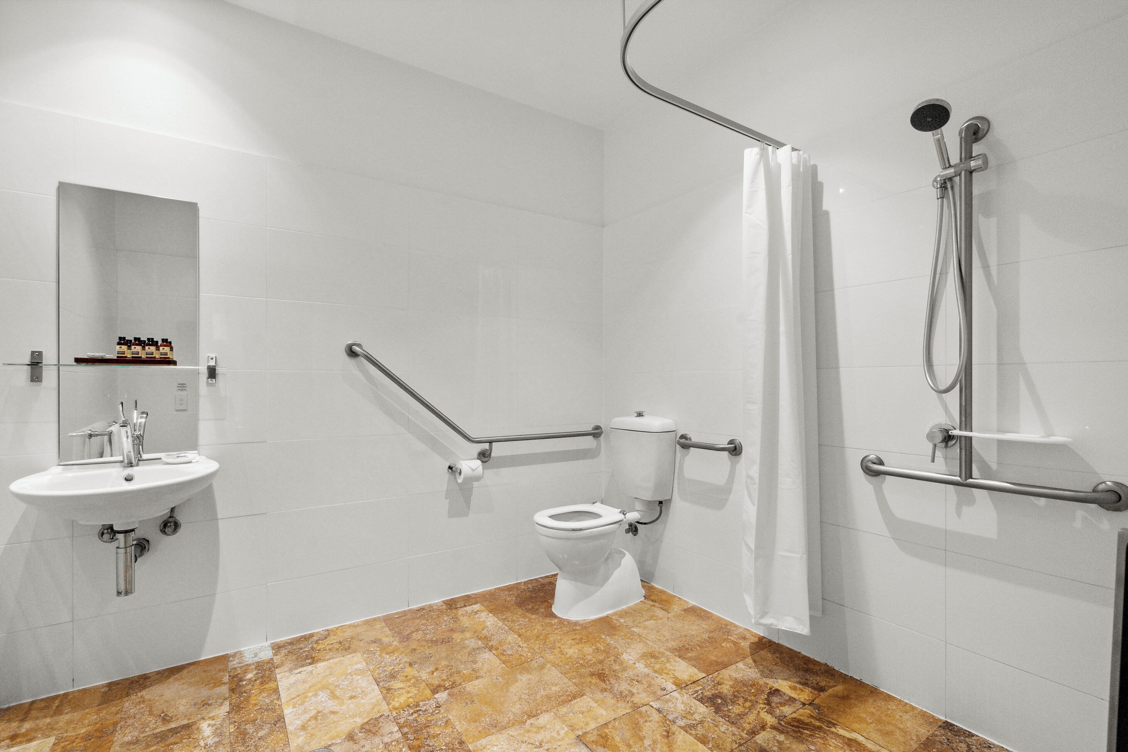 Un baño moderno con lavamanos, inodoro y ducha blancos. Cuenta con barras de apoyo y un espejo.