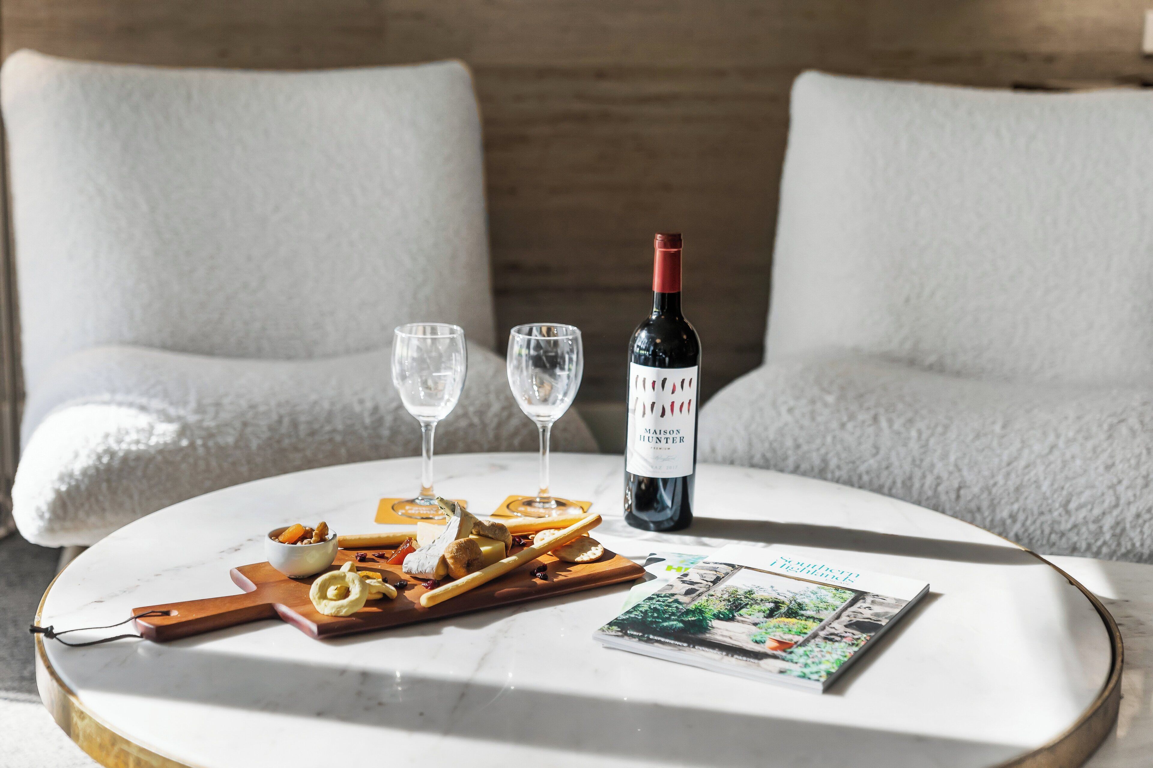 A round marble table with a bottle of wine, two glasses, a magazine, and a wooden board with snacks.
